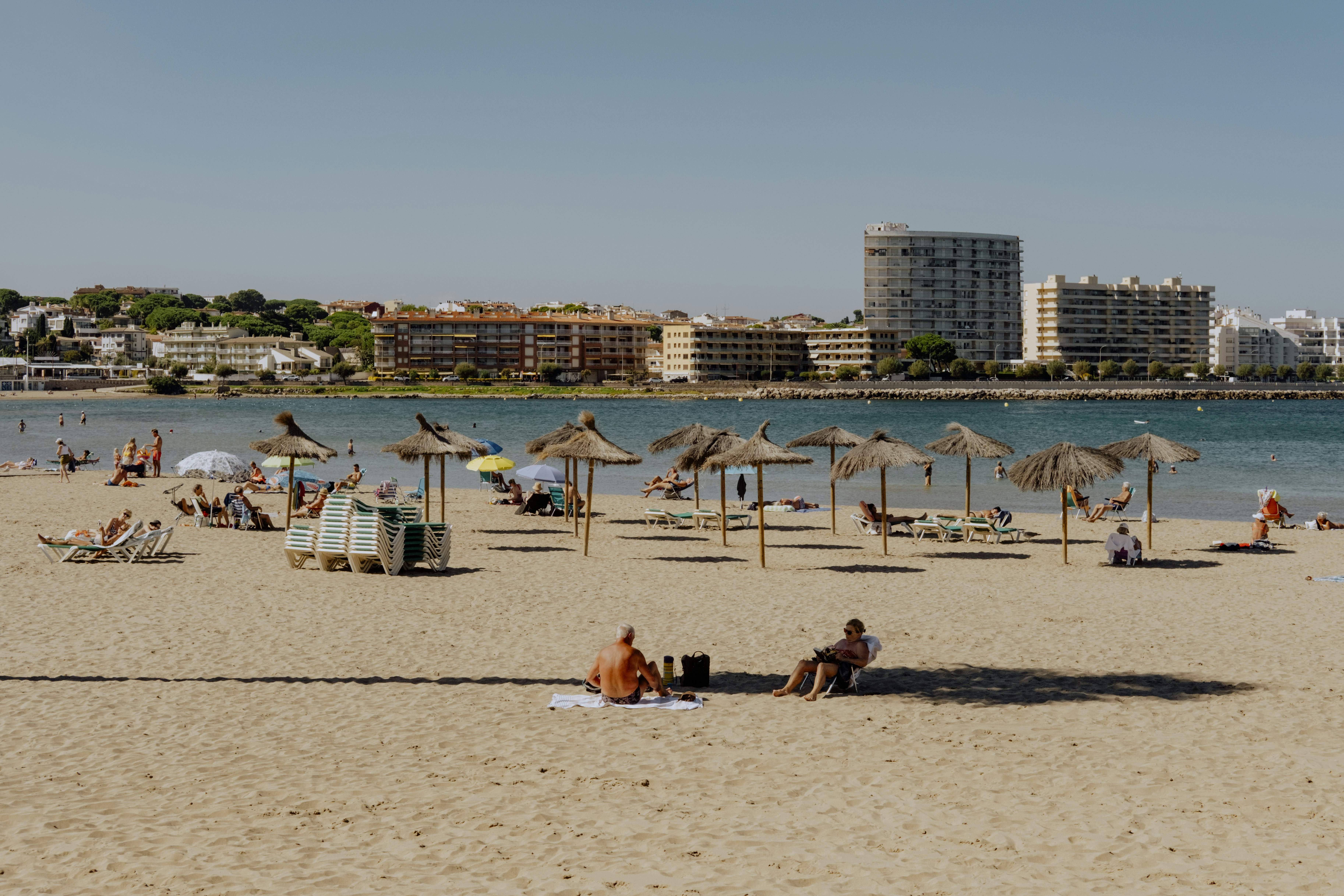 People scattered on a beach with strawlike umbrellas planted in the sand. Low-rise buildings line the coast in the background