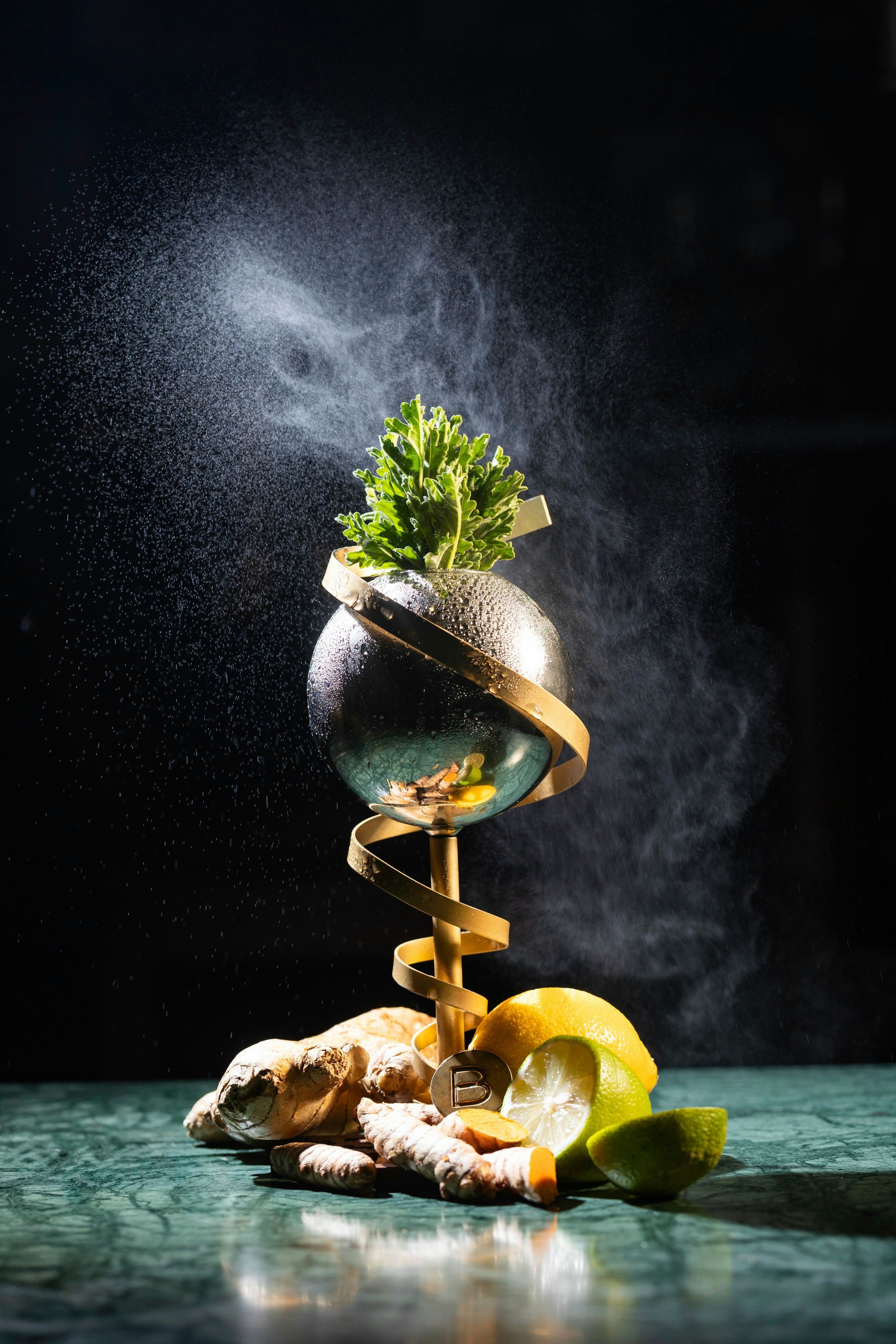 A tall-stemmed drinking vessel with a spherical cup surrounded by fresh cocktail ingredients, including lime, lemon and ginger.