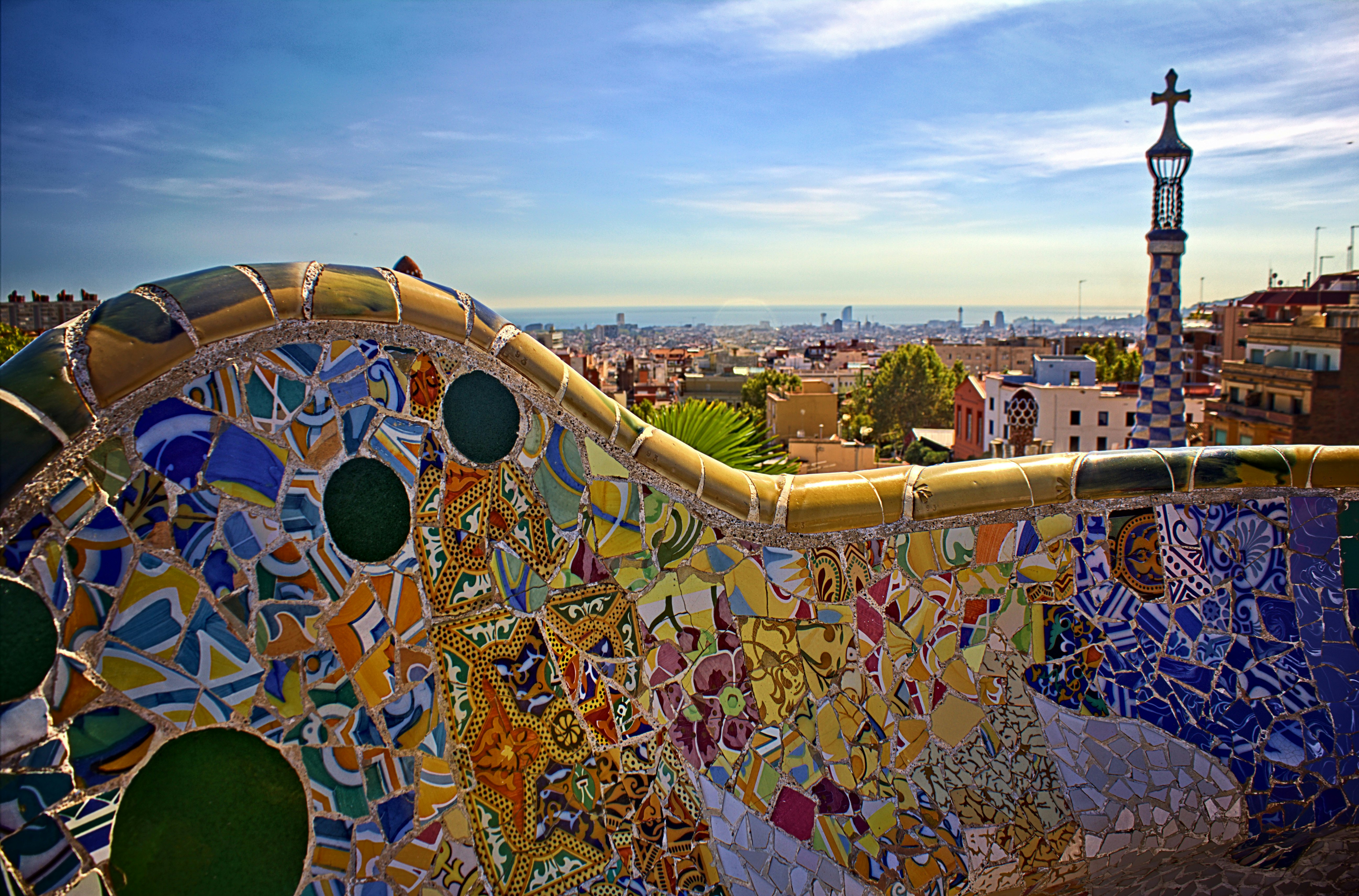 Multicolored mosaic designs on a bench at a park in Barcelona, Spain; a checkerboard tower with a cross is visible in the background.