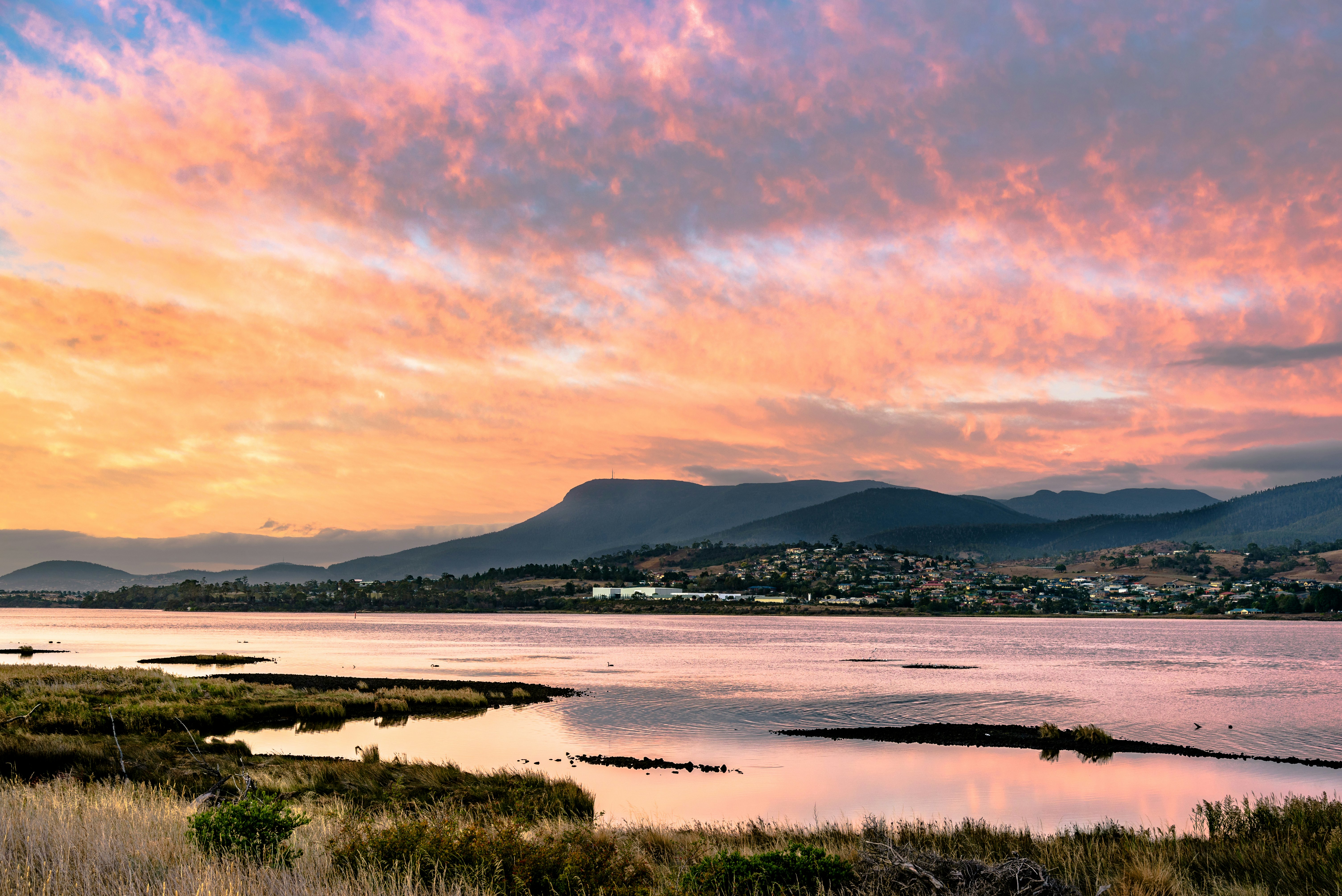 The sunrise casts a pink and orange glow over a wide river flowing near a small hillside settlement.