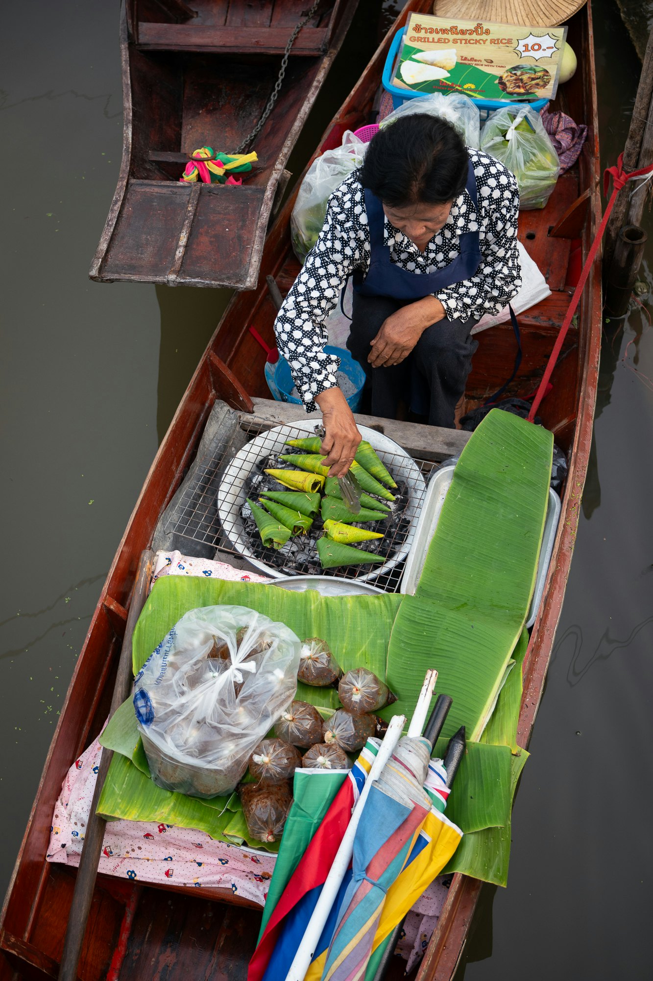 An overhead view of a woman grilling food wrapped in banana leaves on a boat in a floating market.
