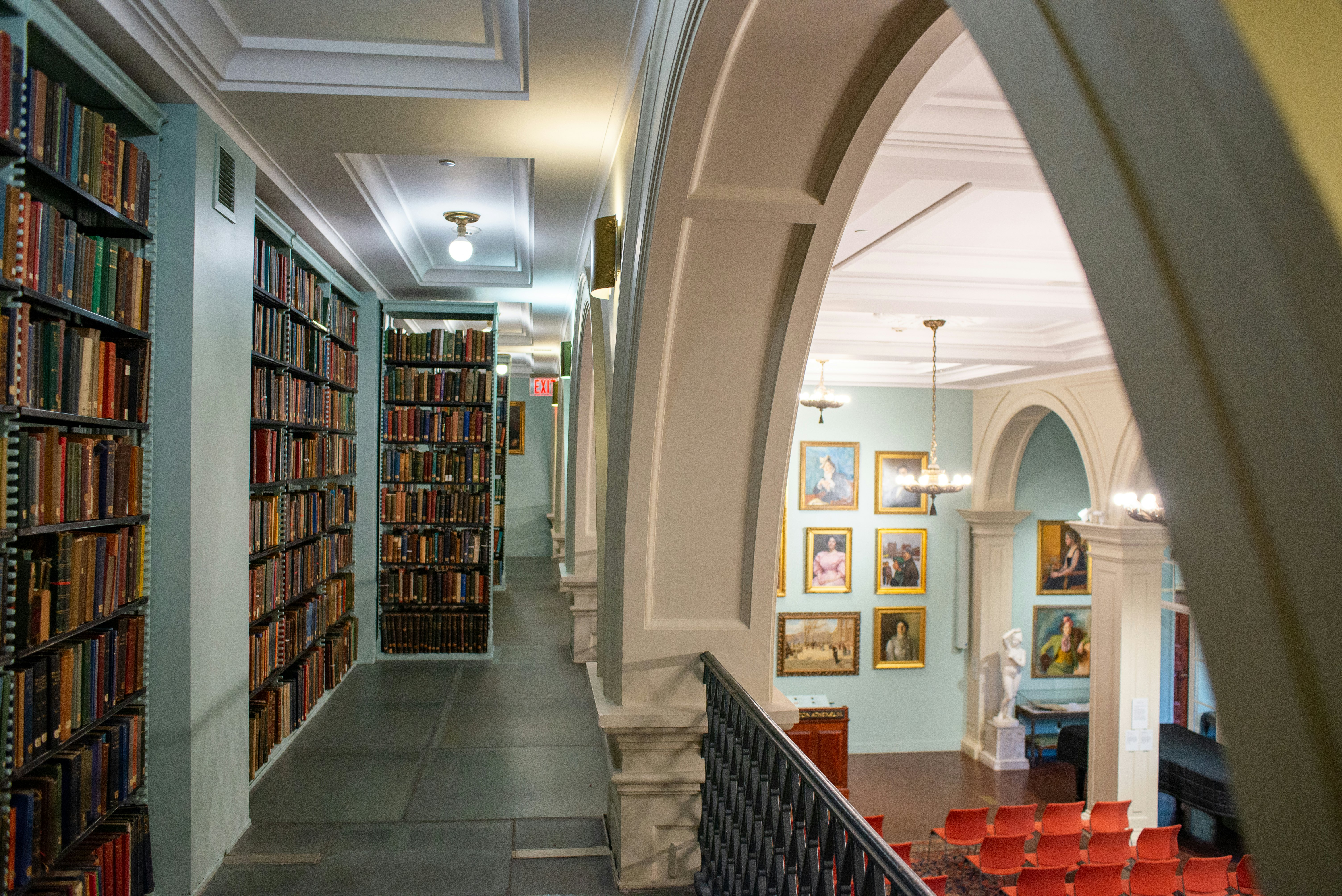 The interior of a historic library, with hallway lined with shelves of books on one side, and a archway and railing overlooking an auditorium space on the other.
