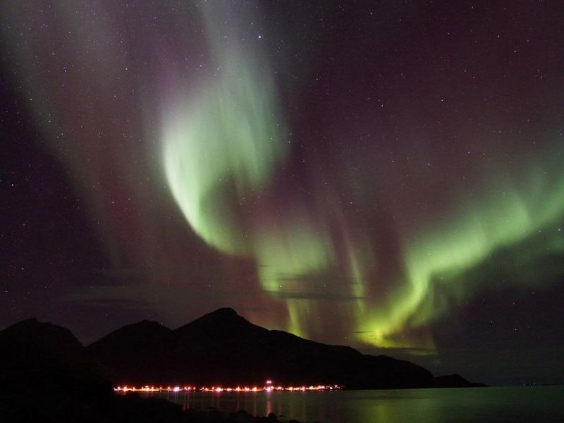 Greenish northern lights in a dark sky over a mountain with lights along the shore.