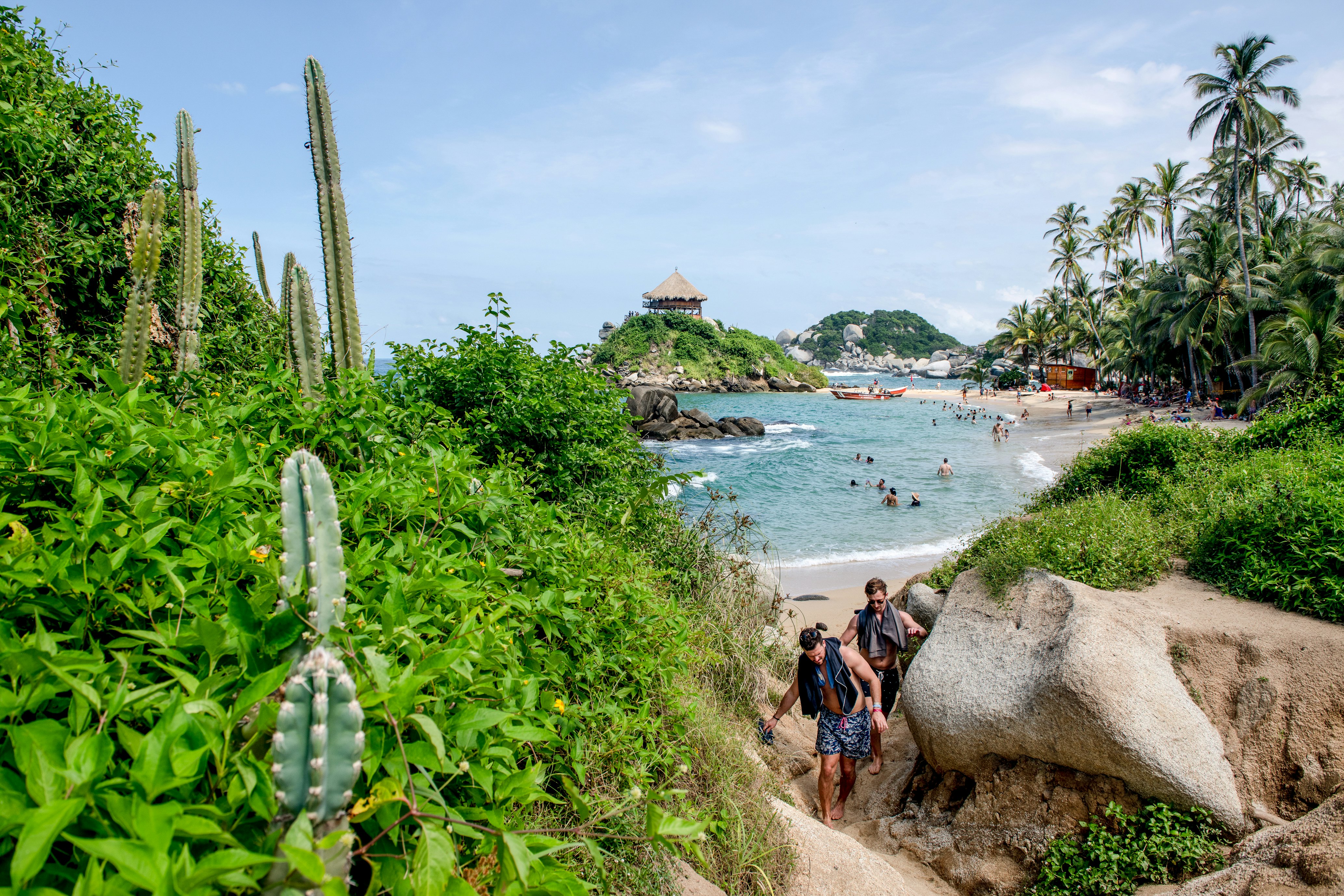 Two people clamber over boulders near a sandy path leading away from a tropical beach on a sunny day.