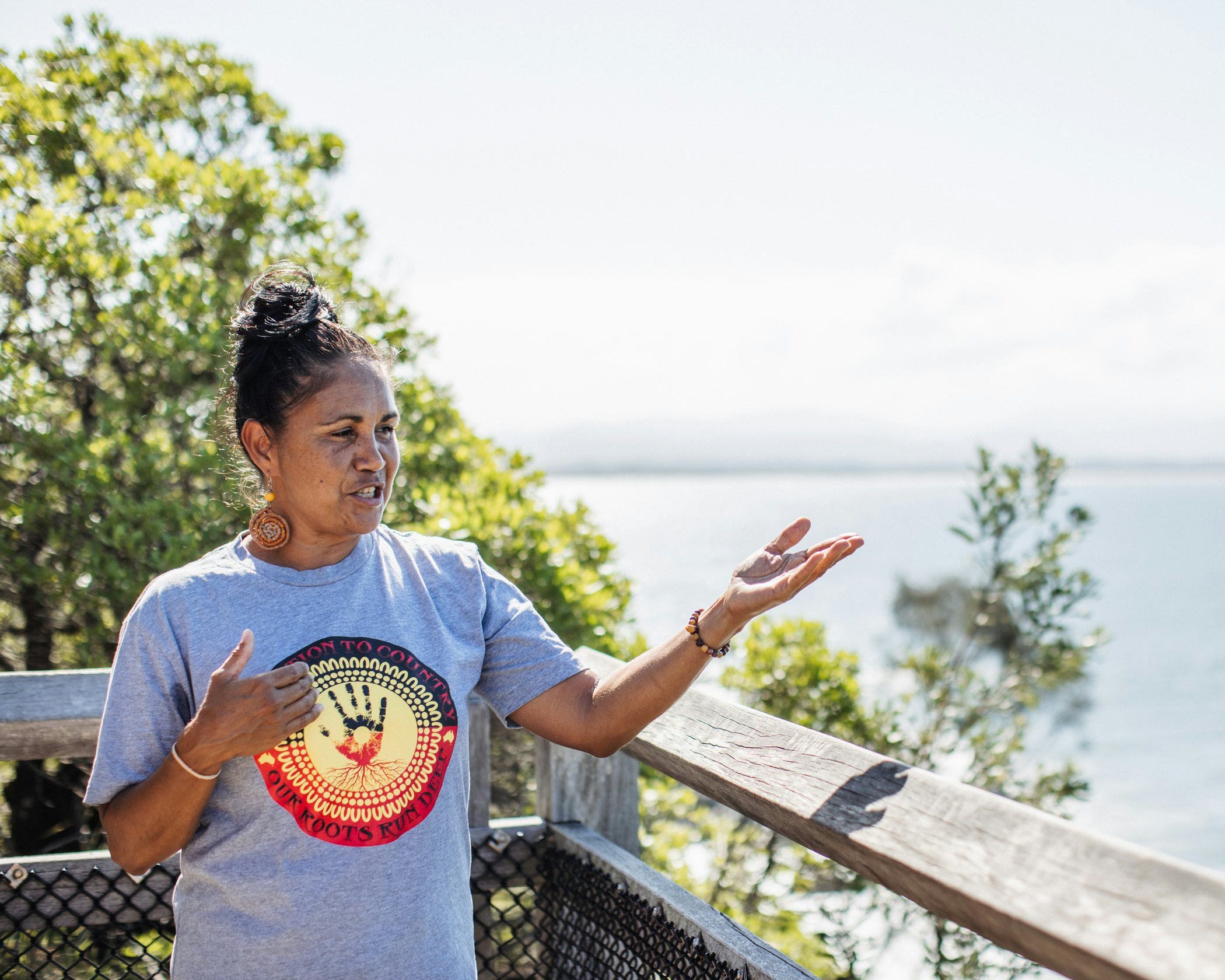 A woman wearing a T-shirt reading "Our roots run deep" gestures from a balcony during an Explore Byron Bay tour