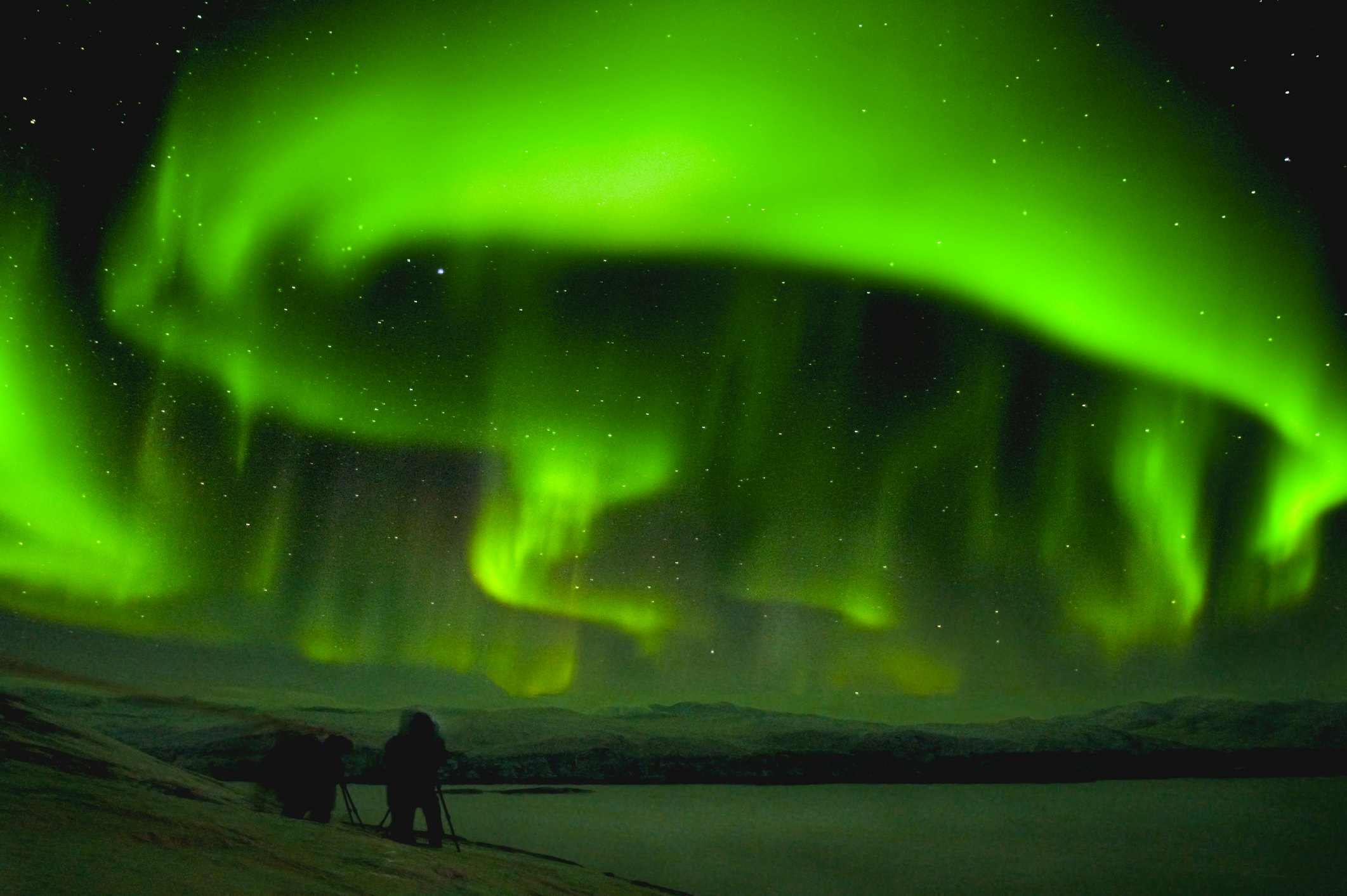 Huge streaks of green northern lights over a dark landscape with the silhouette of a photographer and equipment in the lower left.
