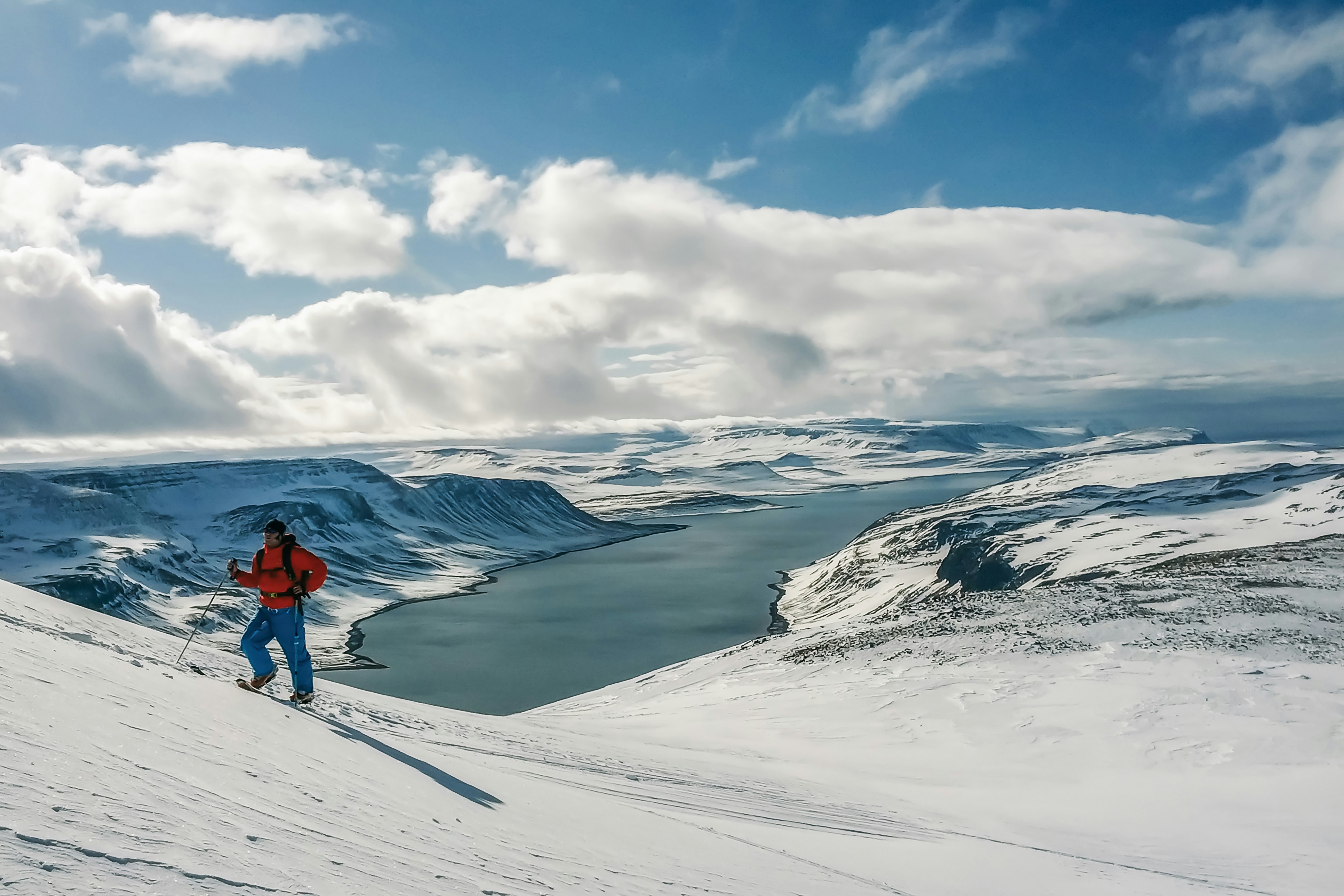 A mountaineer in red snow gear hikes up a snow-covered hill above a fjord.