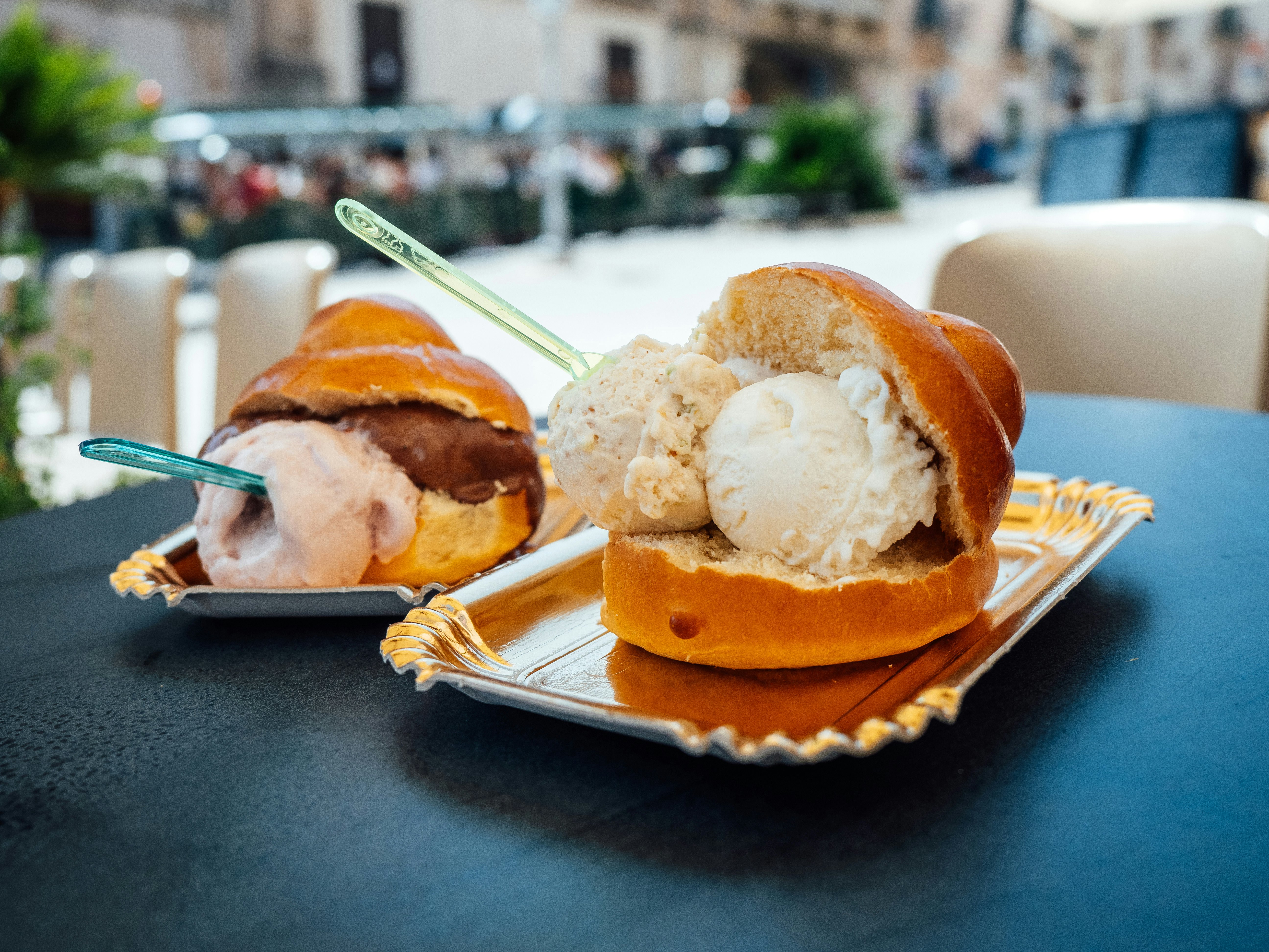 Two brioches stuffed with ice cream sit on disposable plates on the table of an outdoor cafe.