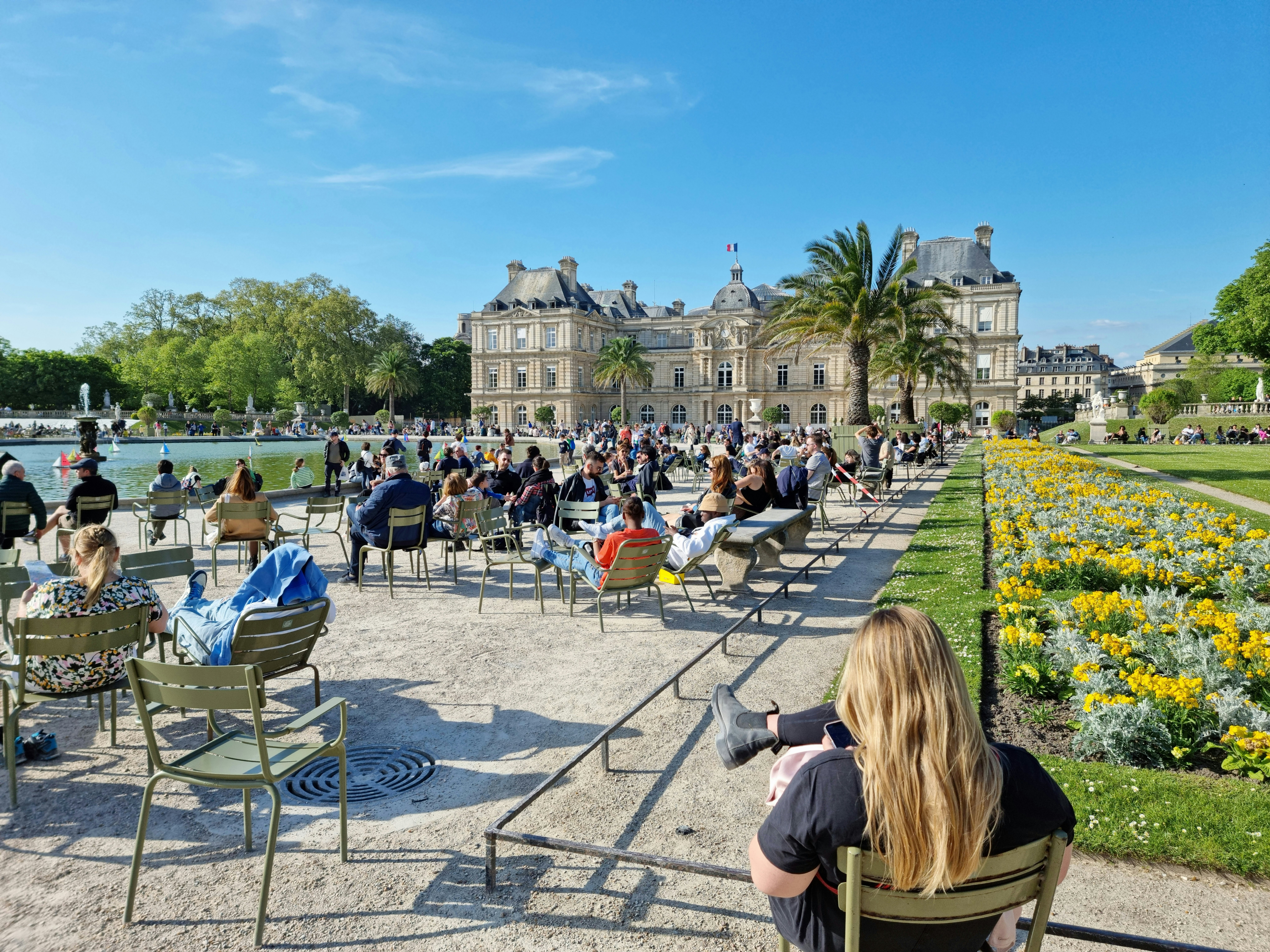 People relax in chairs and loungers on a stretch of gravel pathway set between flower-filled borders and a small artificial lake in a large public park overlooked by a grand mansion.