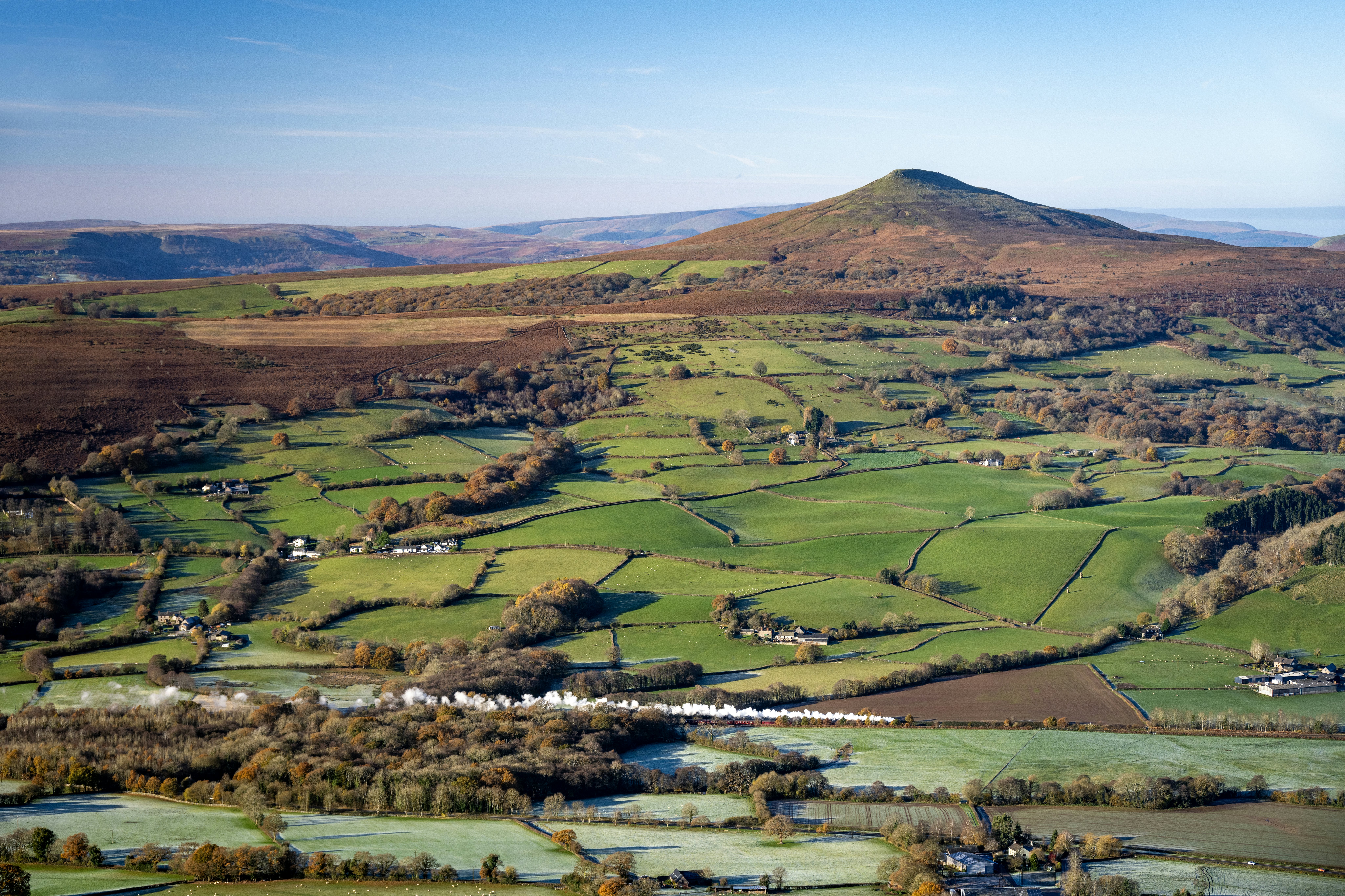A steam train follows a rail line through hilly countryside overlooked by one prominent hillock.