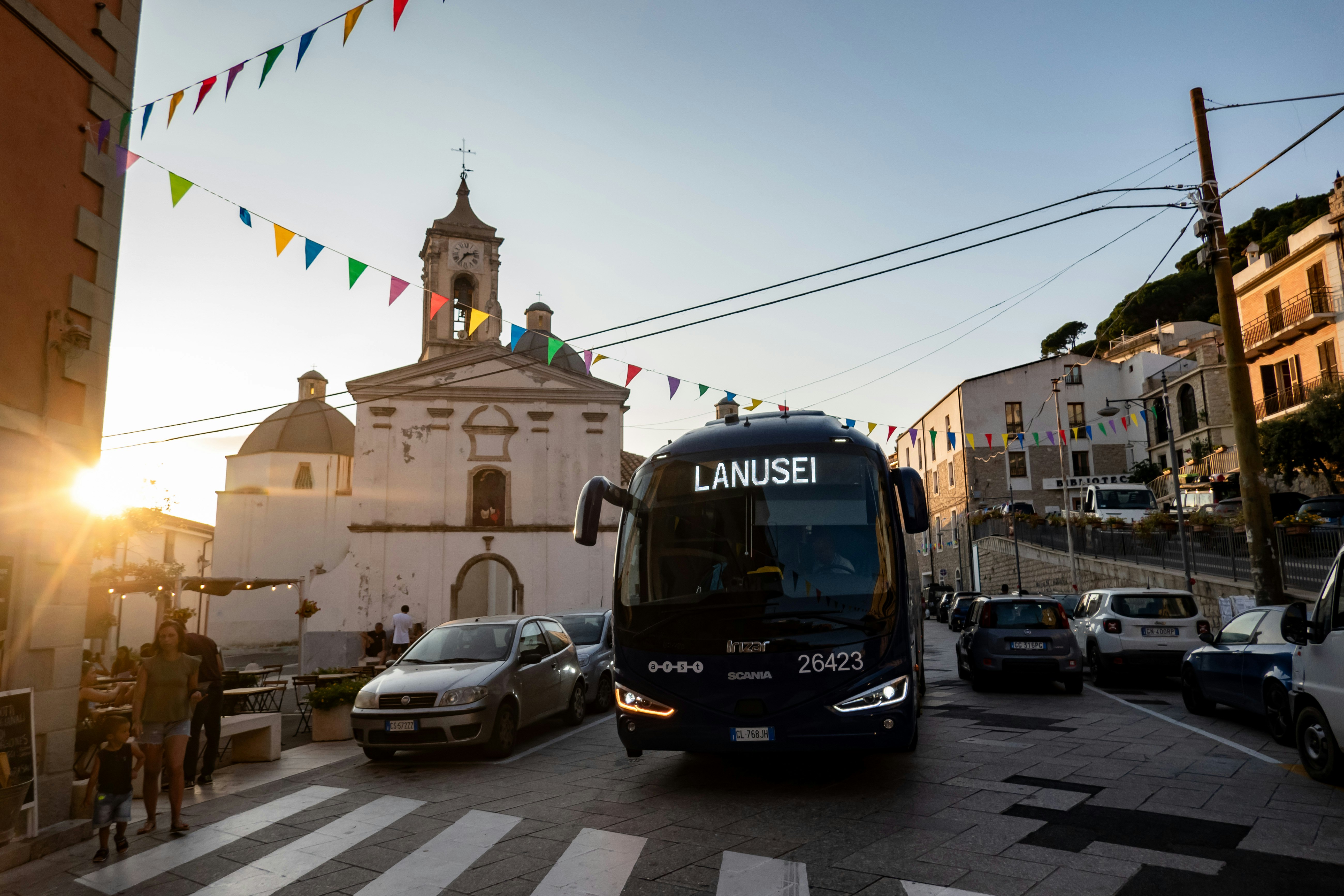 A bus passes through a village square at sunset, with a church to one side and flags hanging over the street.