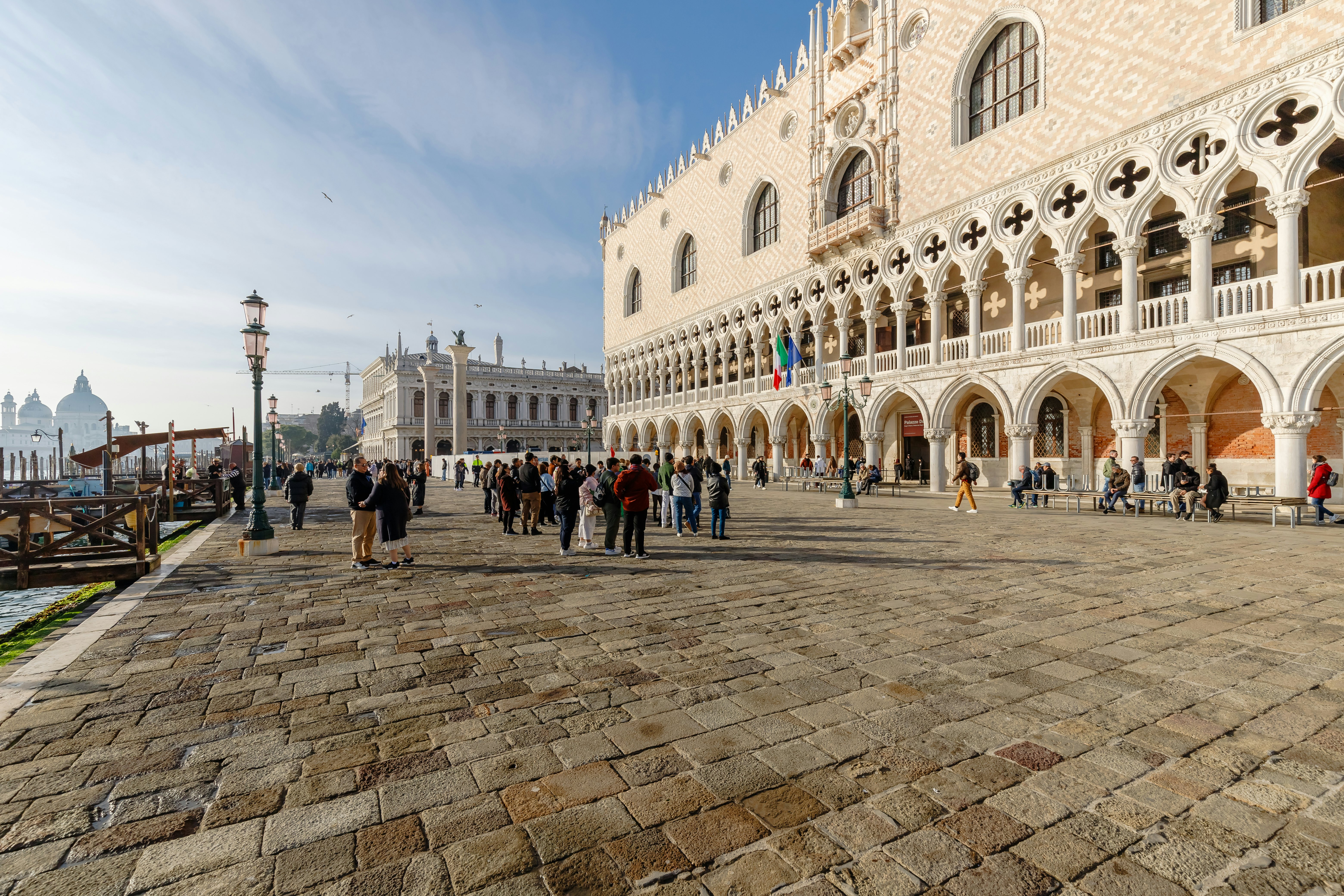 Groups of people outside a grand building with arches in its facade at the edge of a canal.