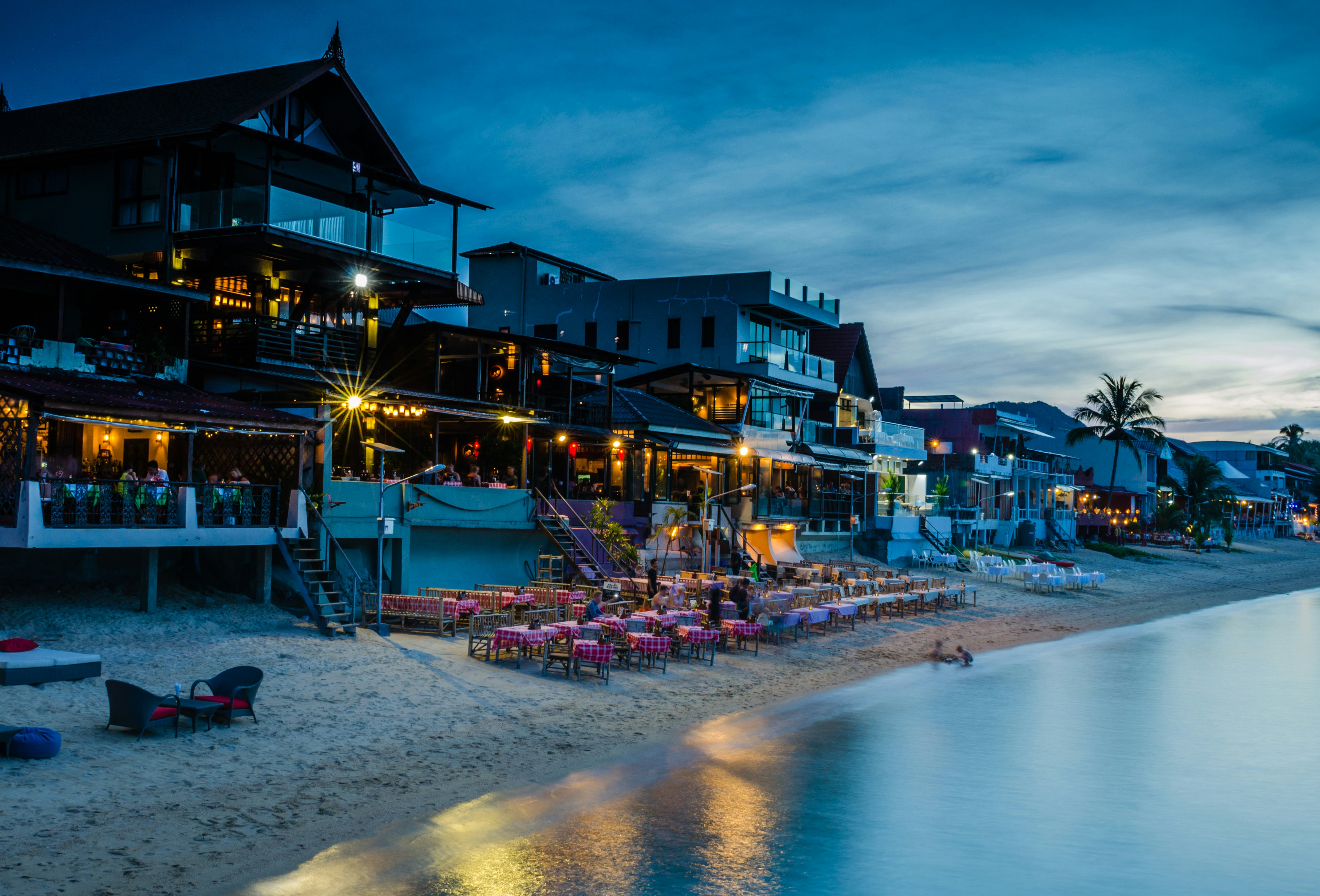 Tables set out on the beach backed by restaurants as twilight falls, lights come on and diners come out for dinner.