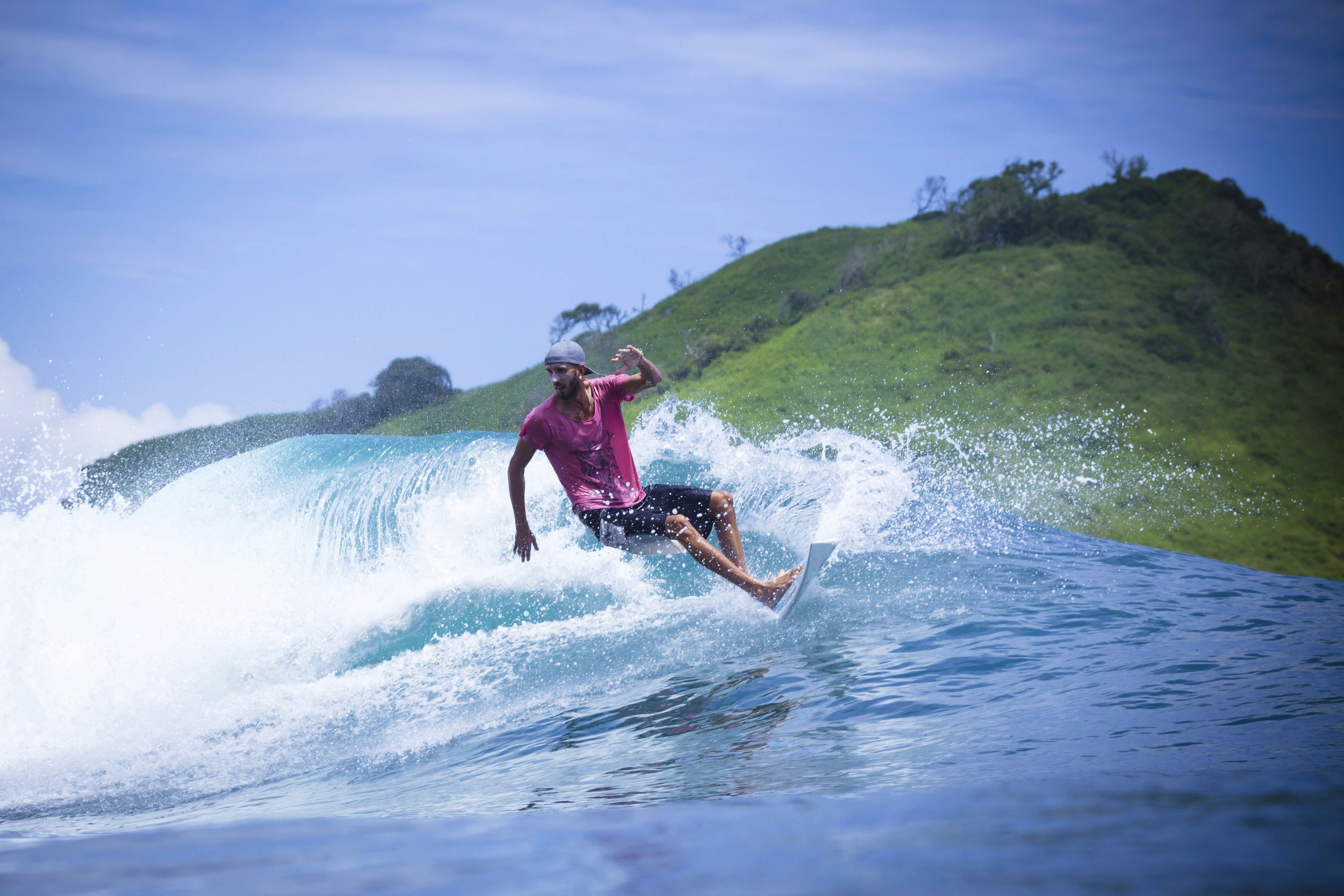 A surfer wearing a pink shirt and a blue cap rides a surfboard with a wave crashing behind him.