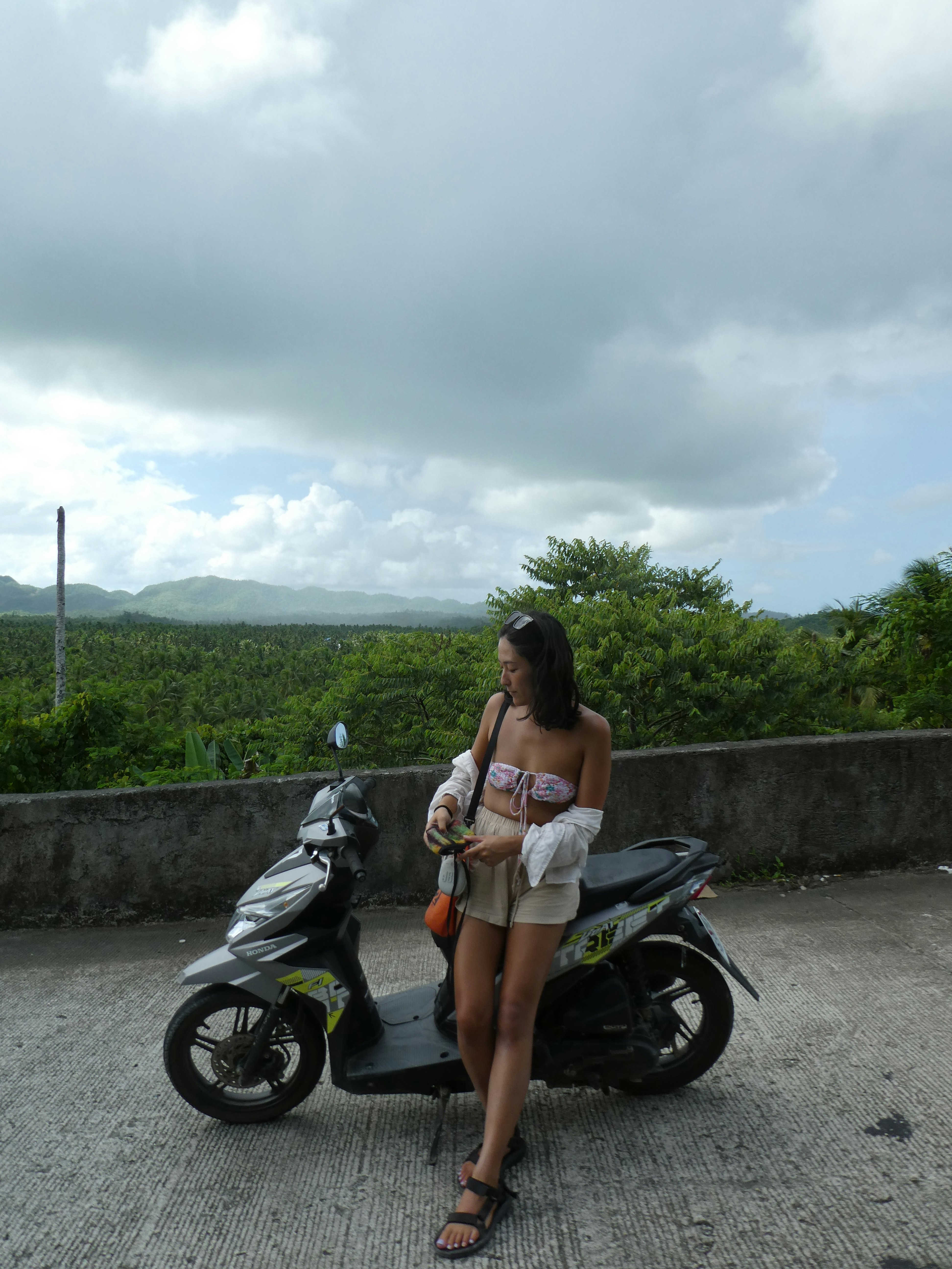 A woman standing next to a motorcycle at the edge of the road with palm trees in the distance.