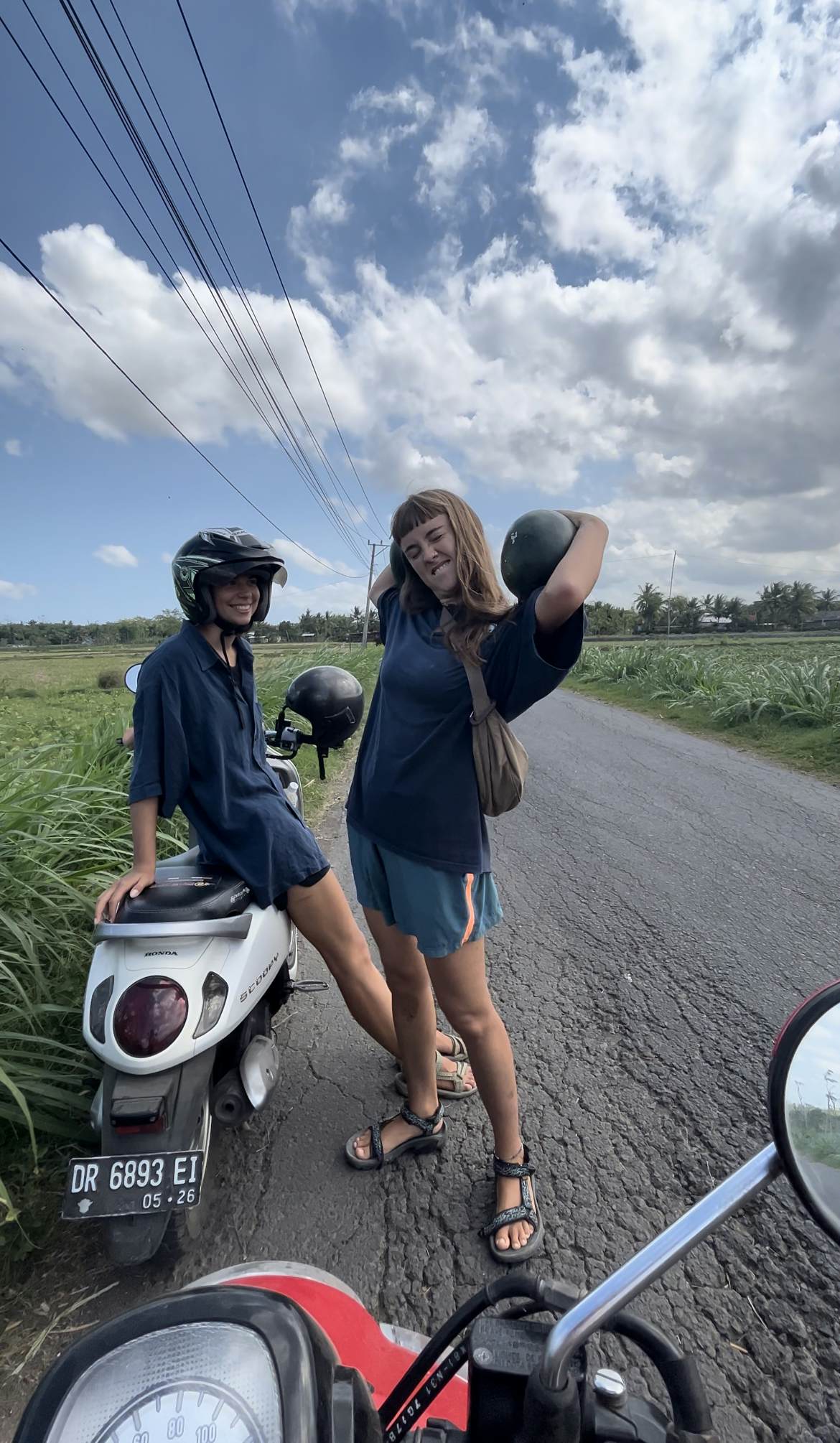 Two women holding watermelons stand next to a motorbike. 