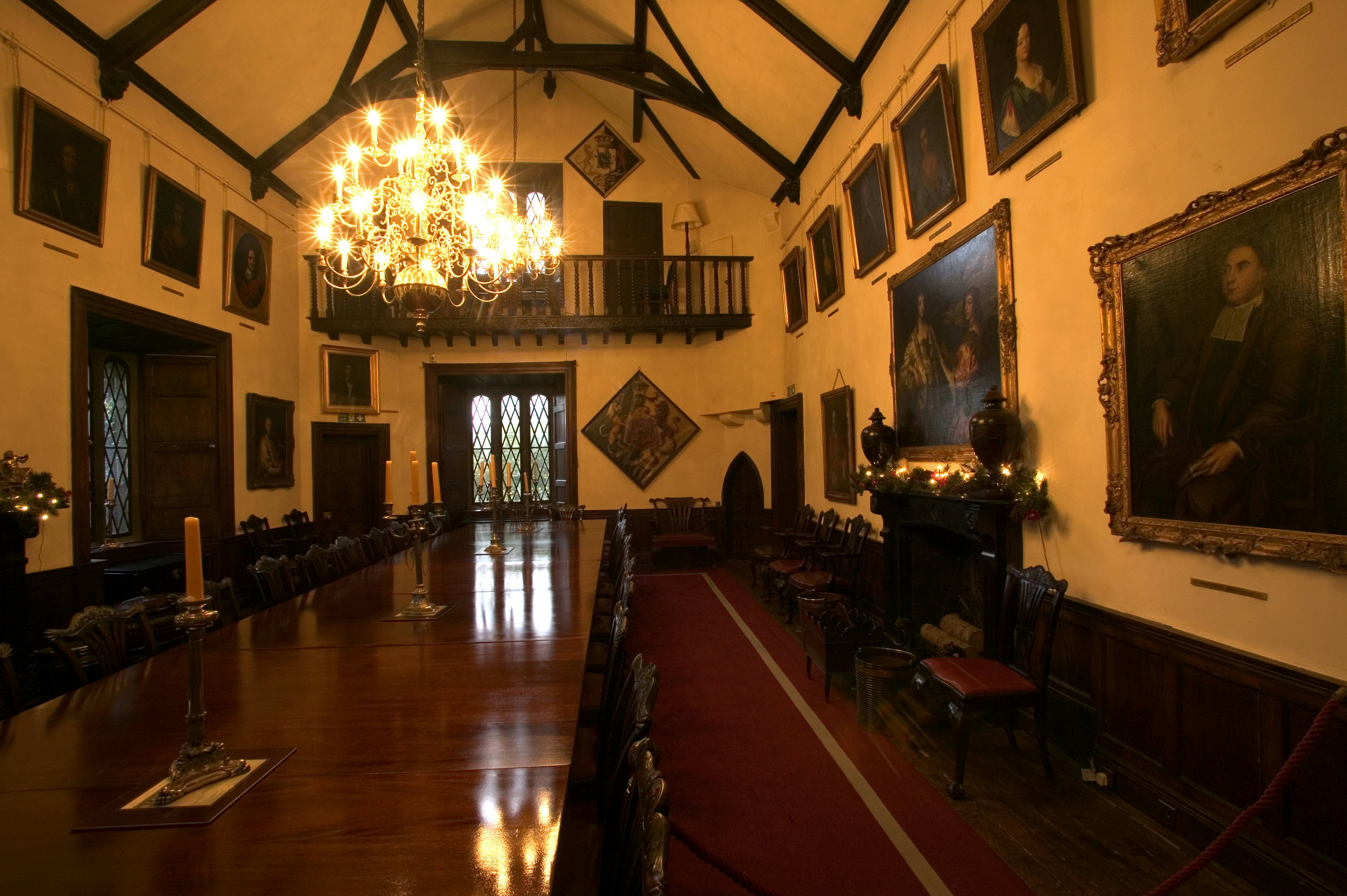 Room with a long, polished dark wood dining table lined with chairs. A chandelier hangs above it, and large painted portraits line the walls.