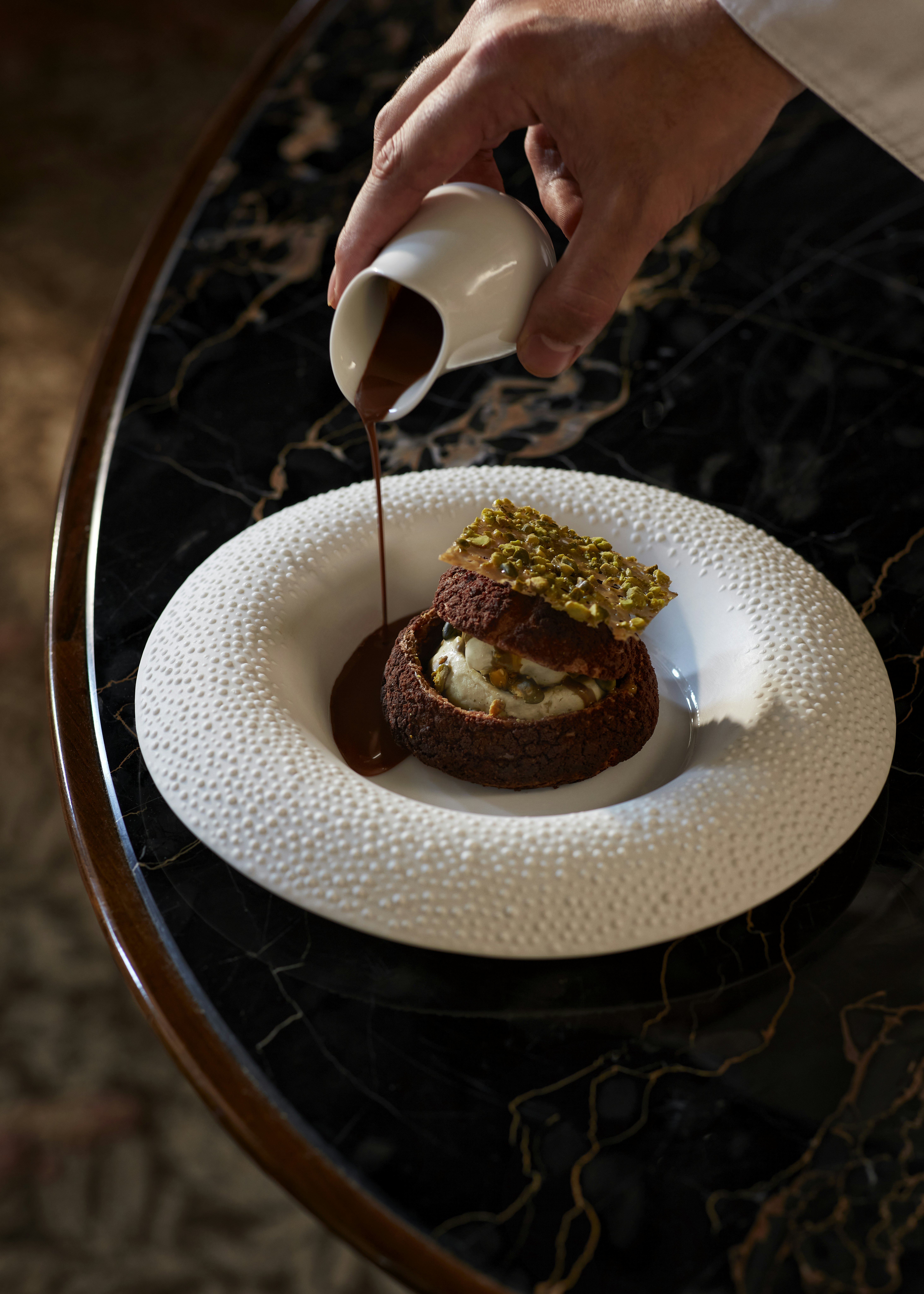 A hand pours chocolate sauce from a small white jug over a delicately presented baked dessert with a pistachio-covered crisp on top.