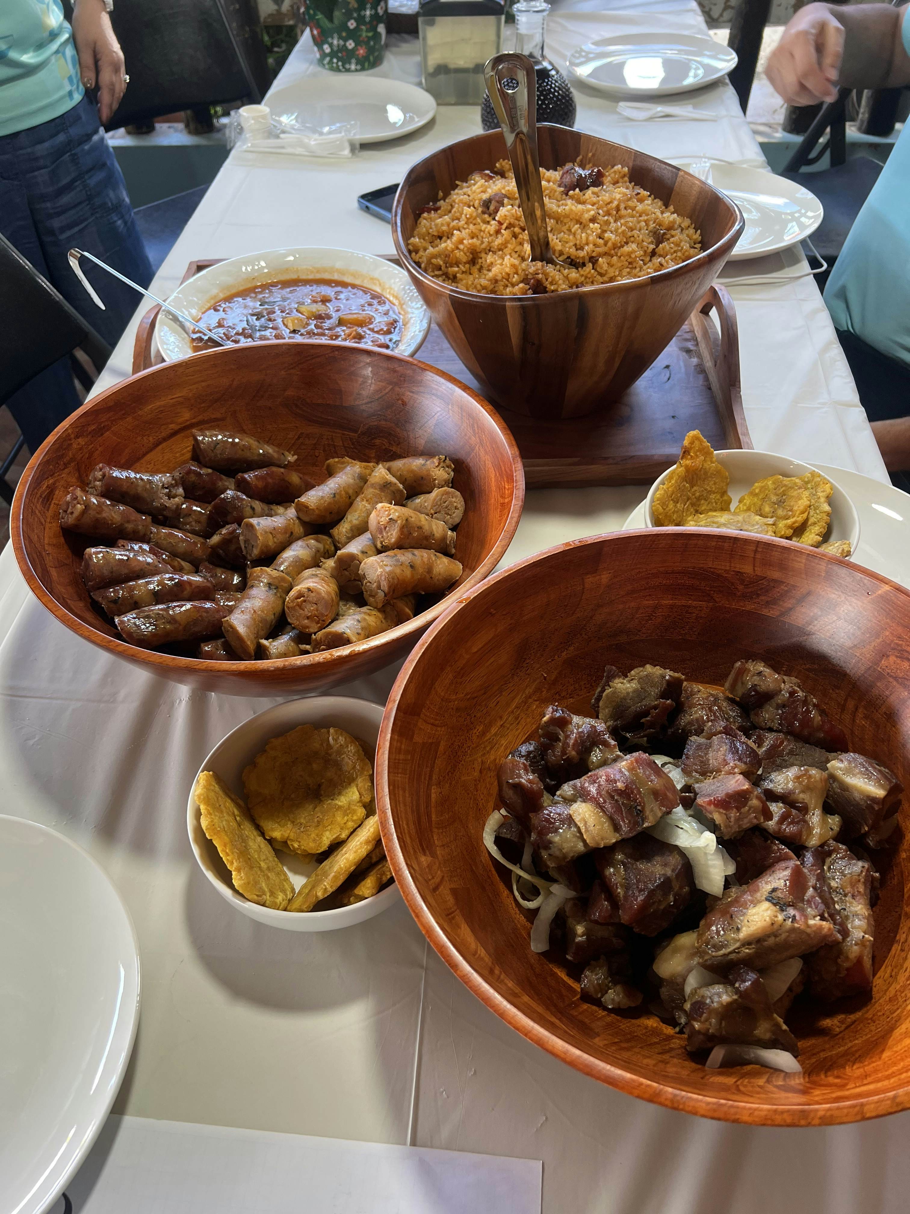 A table set with large wooden bowls filled with sausages, roasted pork, and rice and beans, and smaller bowls with tostones.