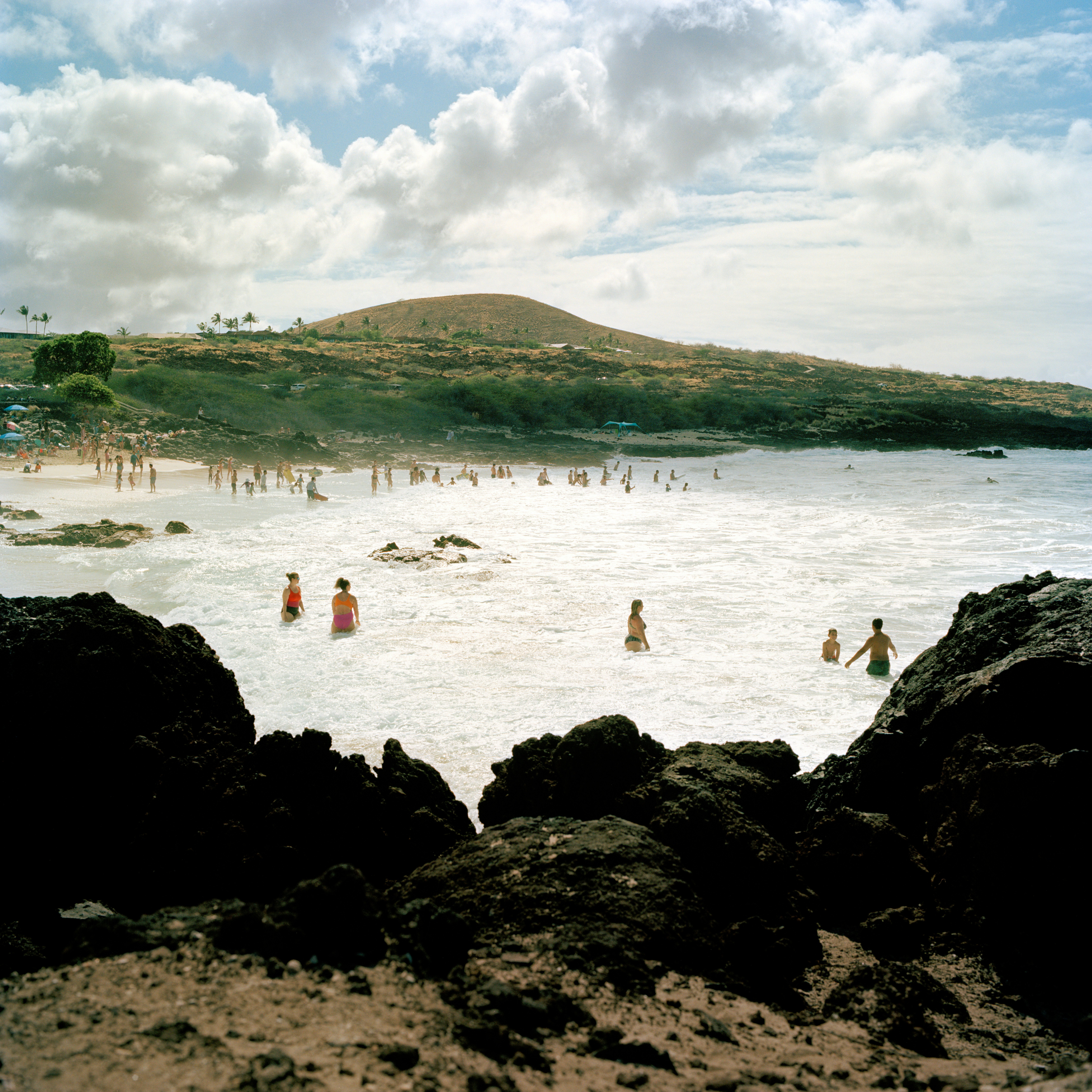 People bathe in the surf on a sandy beach framed by rocks on Hawai'i, with hills behind.