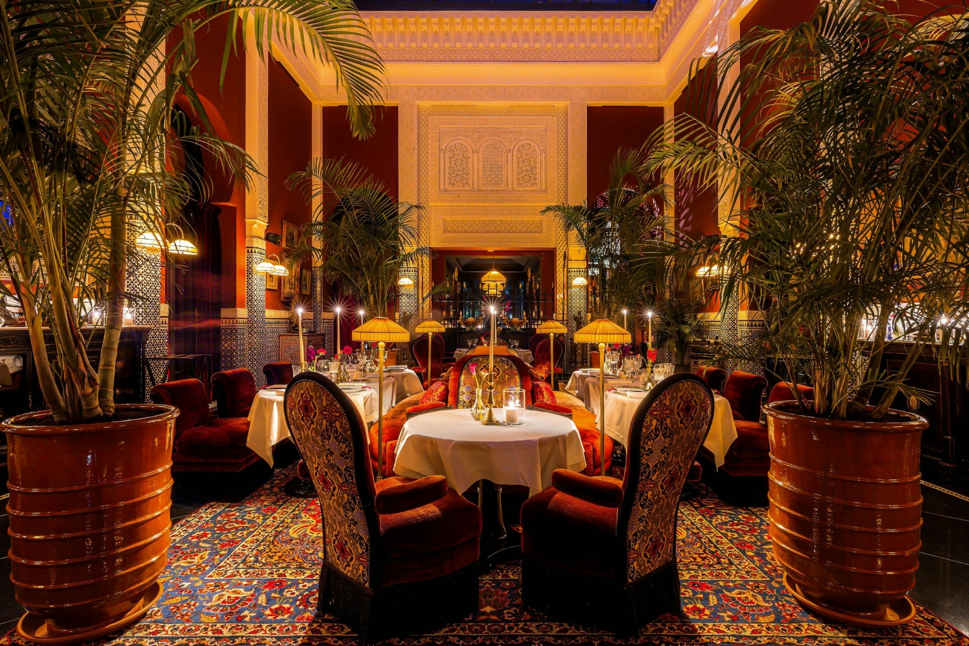A restaurant with patterned rugs and chairs, low lamps and plants in large pots.