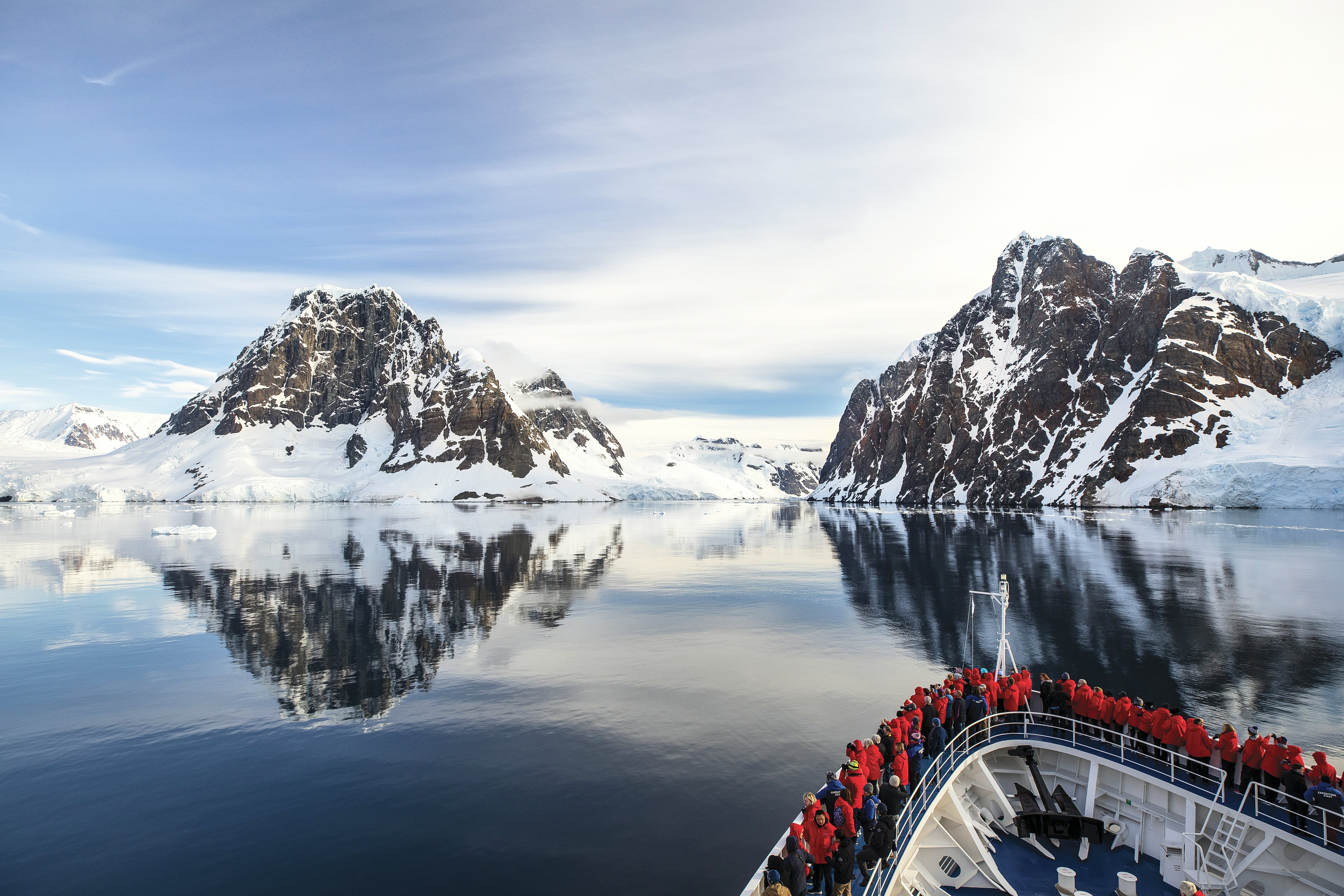 Passengers aboard an expedition ship gather around the bow to look at the surrounding mountains covered in snow in an antarctic ocean.
