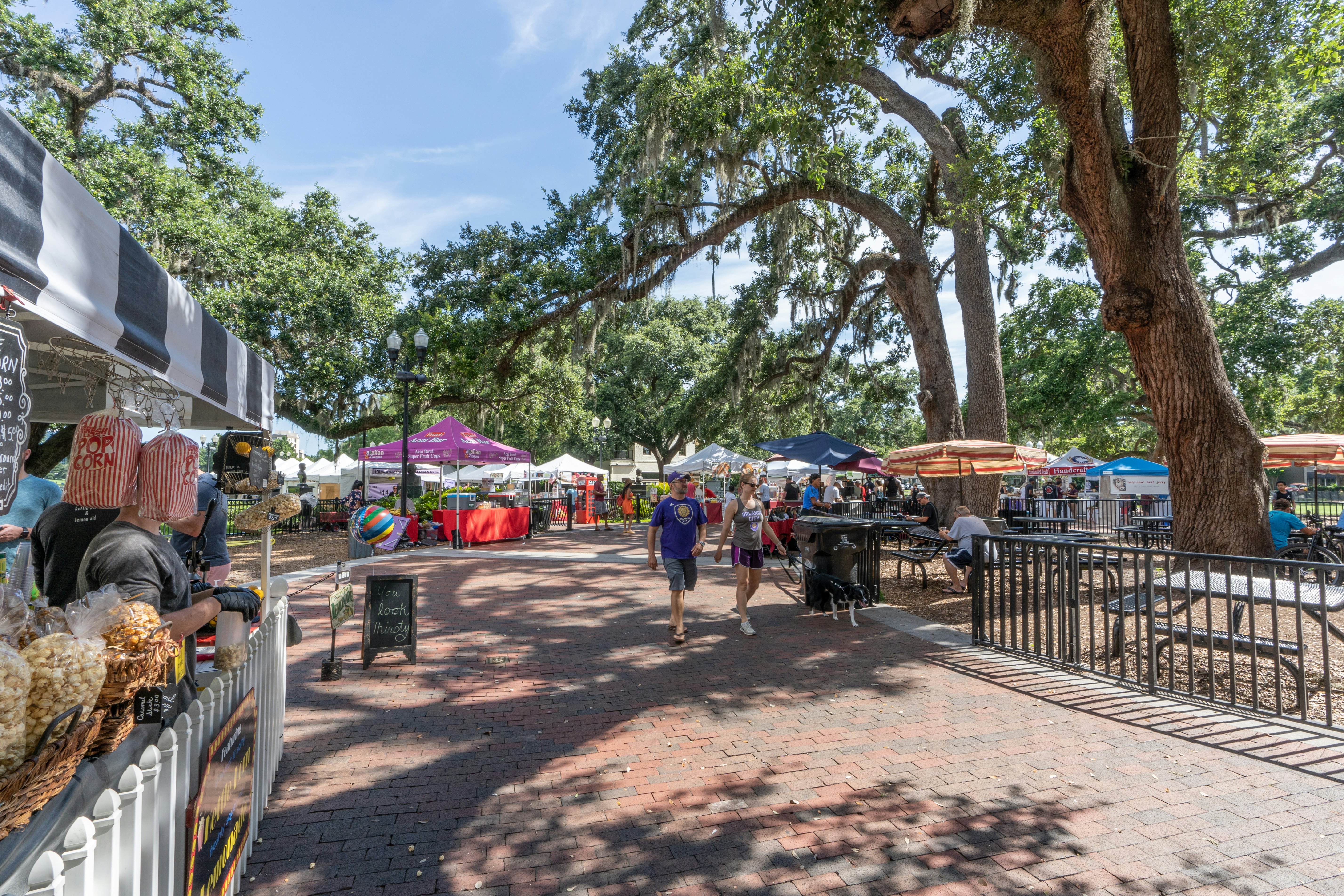 A cluster of booths with colorful umbrellas along a brick walkway under trees with moss hanging from them