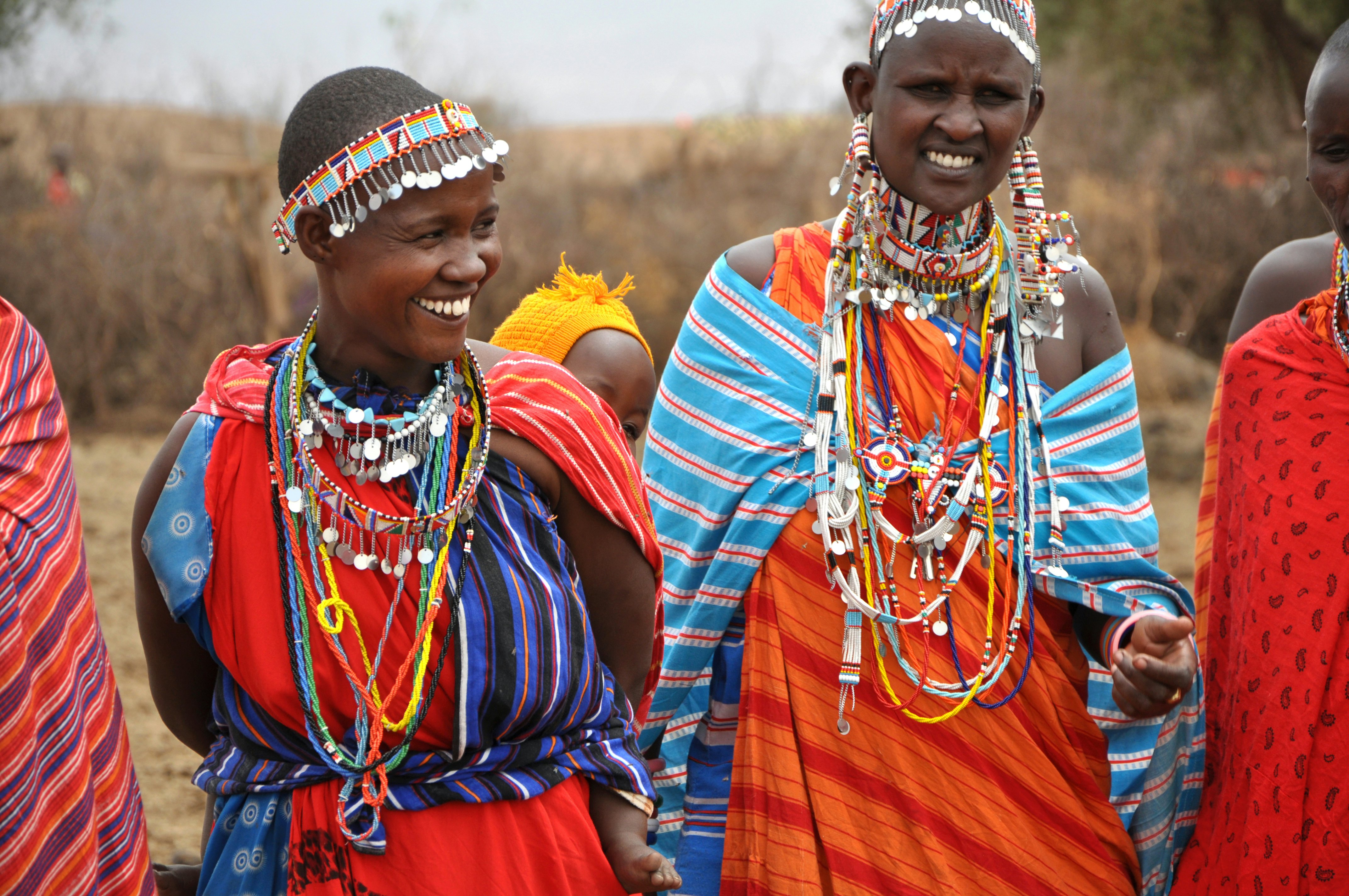Women dressed in brightly colored fabrics and jewelry smile as they pose outside.