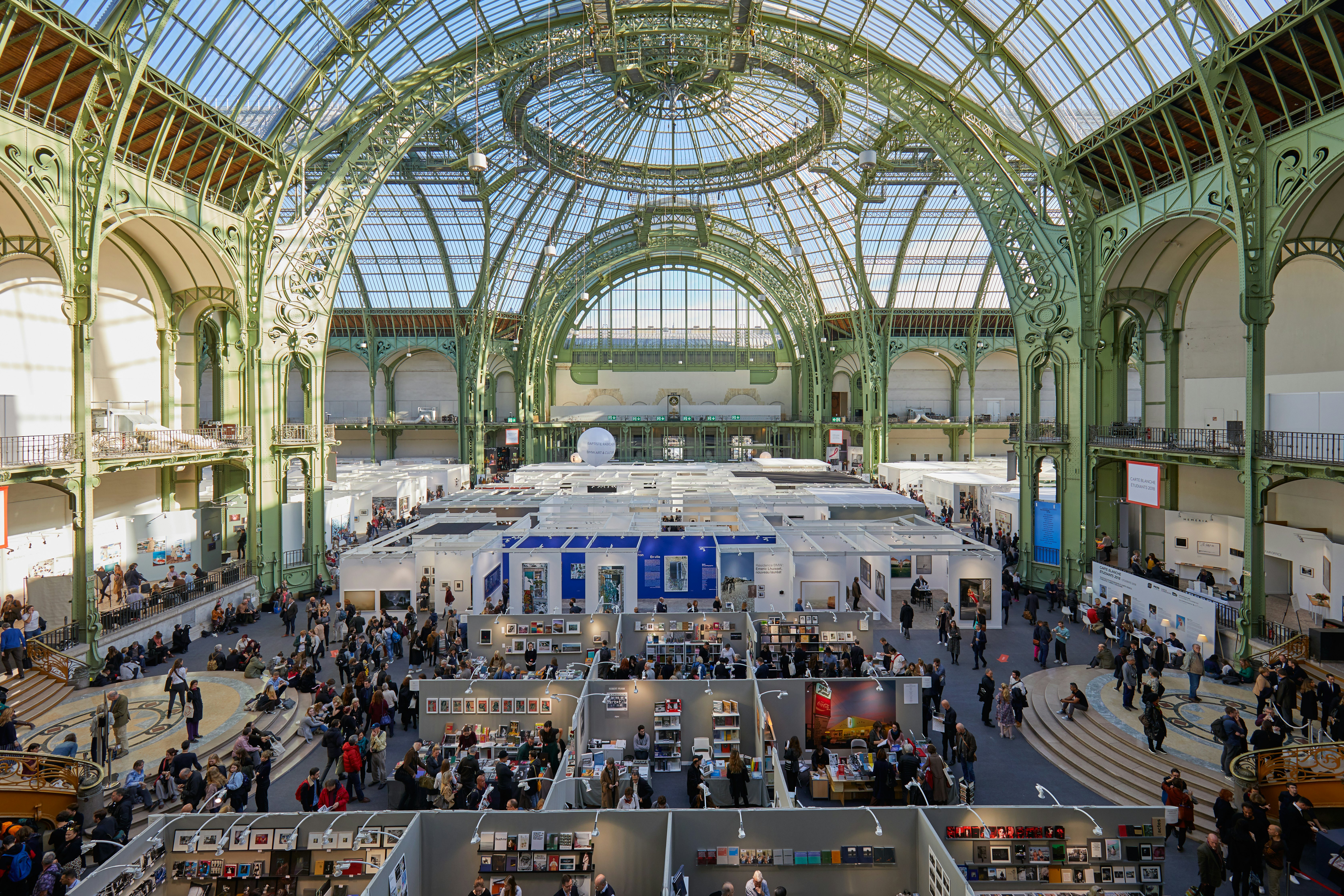 People gather for an exhibition in the main hall of the Grand Palais, featuring a striking iron, steel, and glass barrel-vaulted roof.