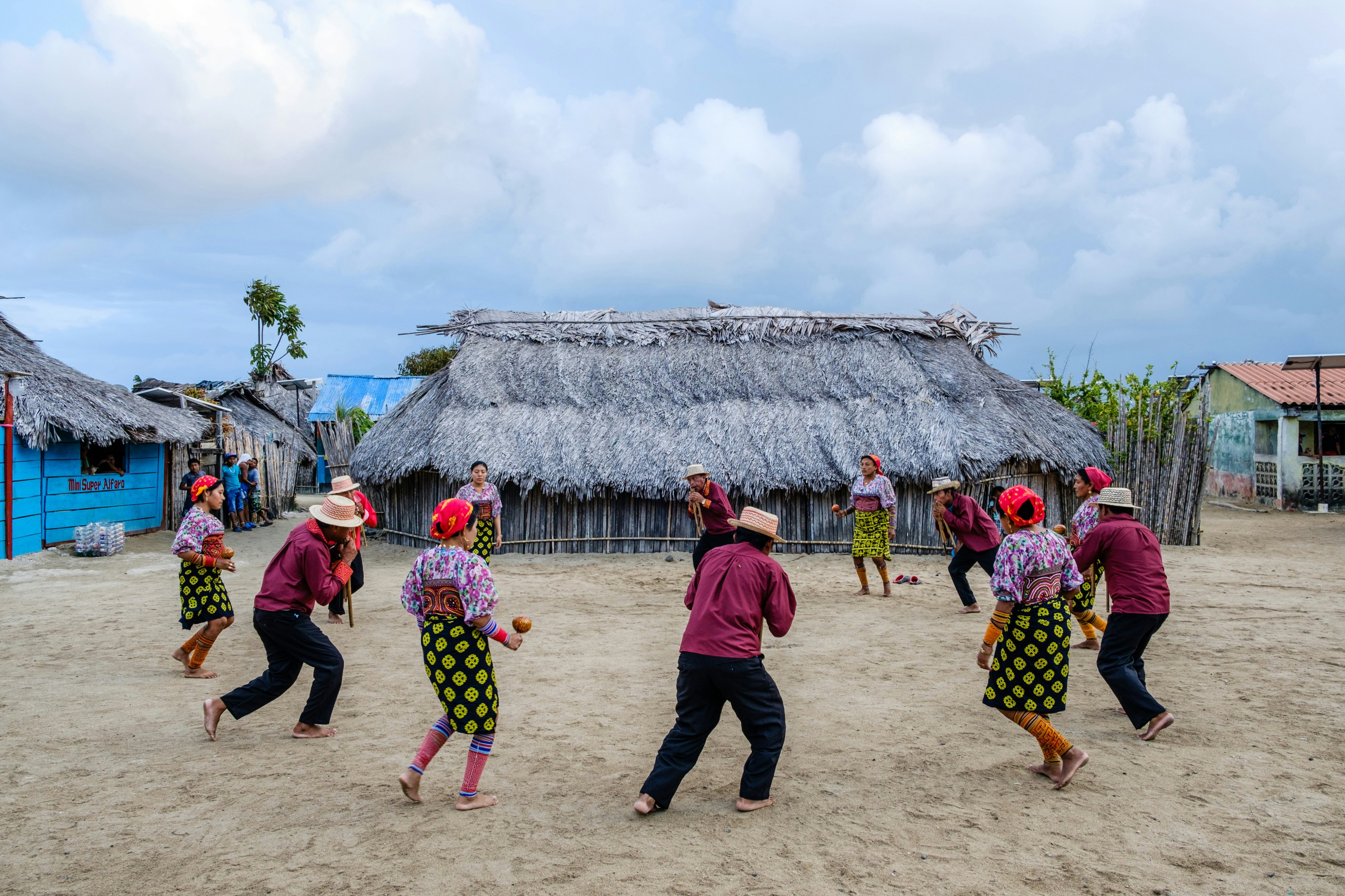 People in brightly colored clothing dance in a circle on the sand in front of a thatch roof building.