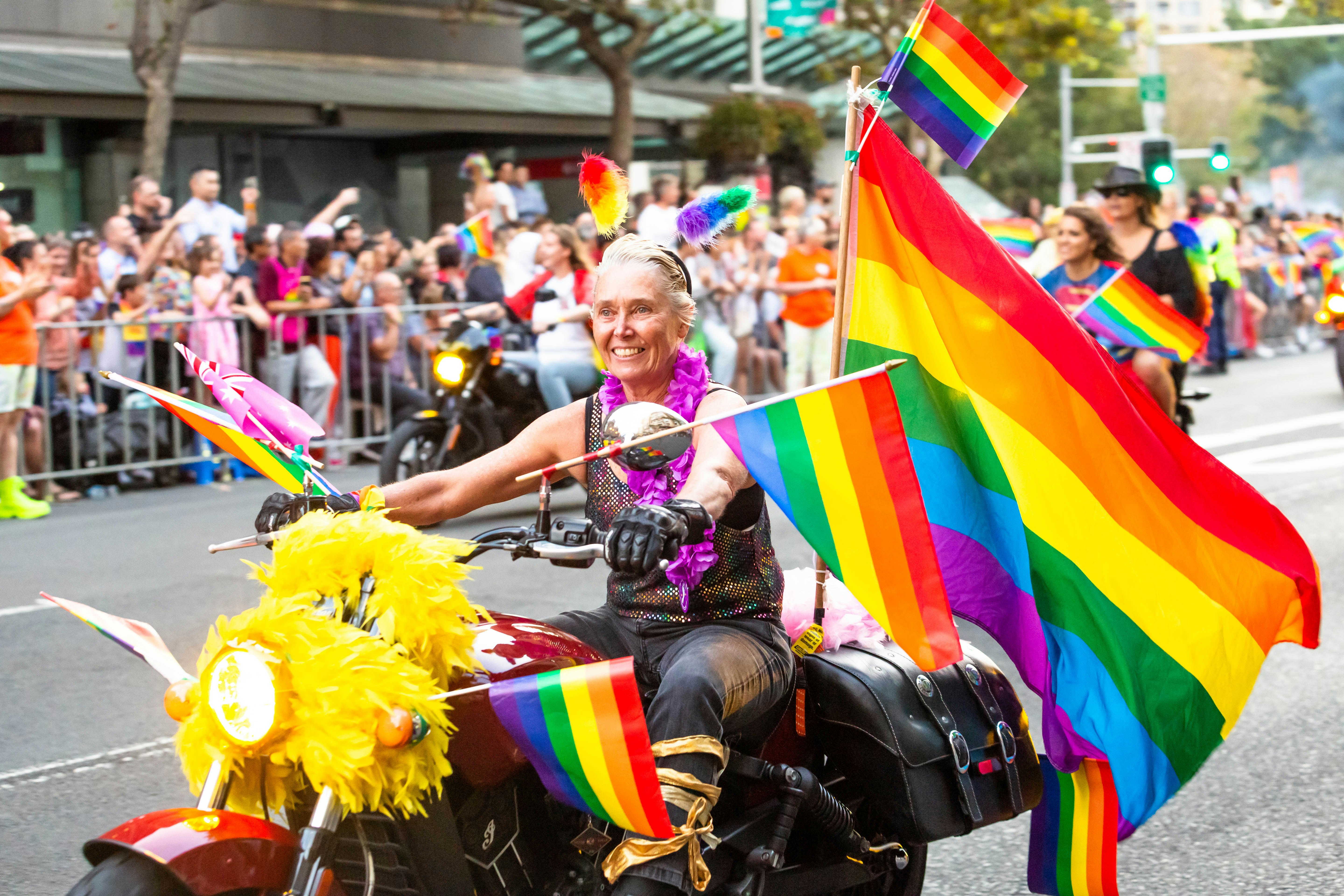 A person in leather on a red motorcycle decorated with rainbow flags of various sizes participating in a street parade