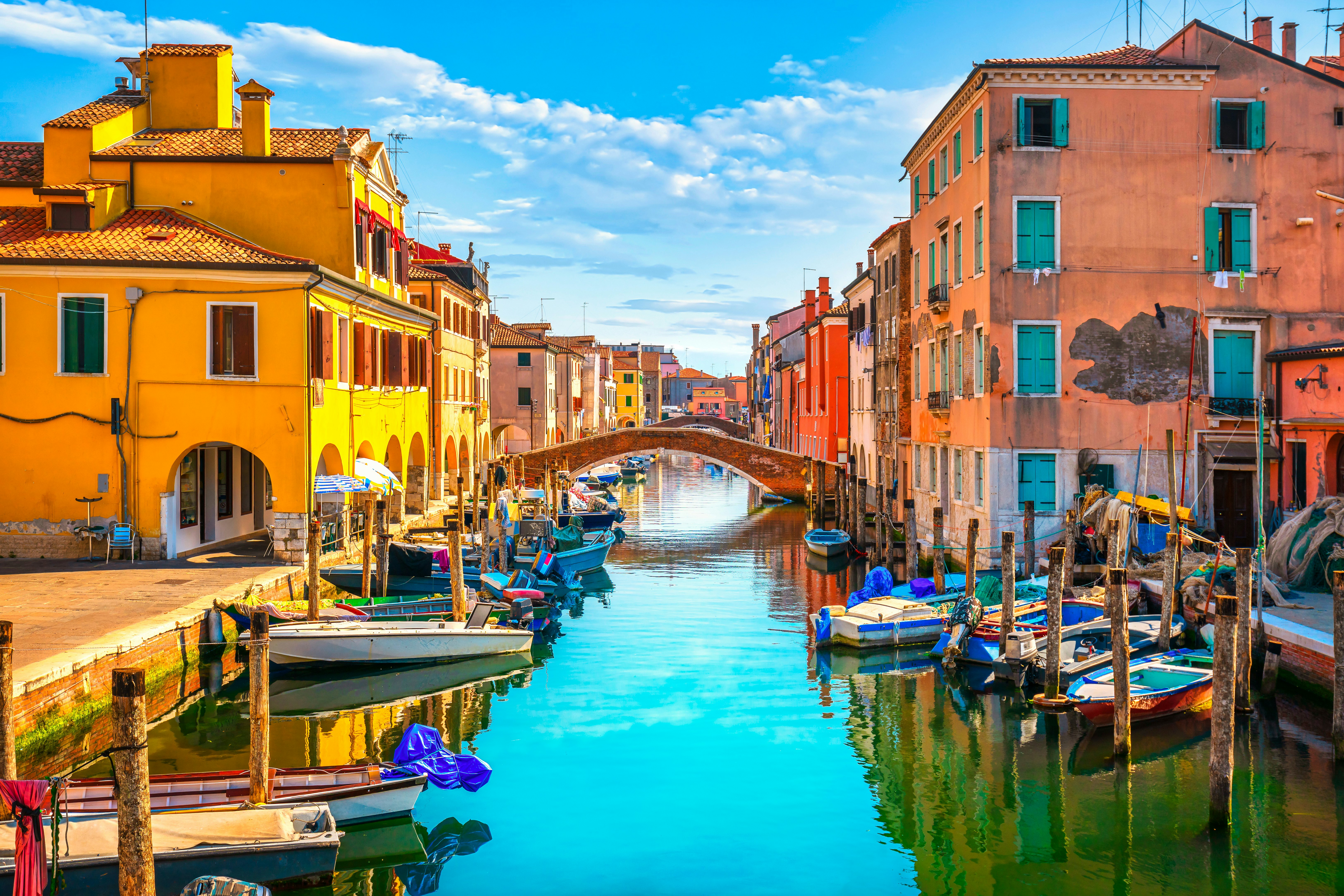 A bridge crosses a canal in Italy, with boats in the water in the foreground; colorful buildings line either side of the canal.