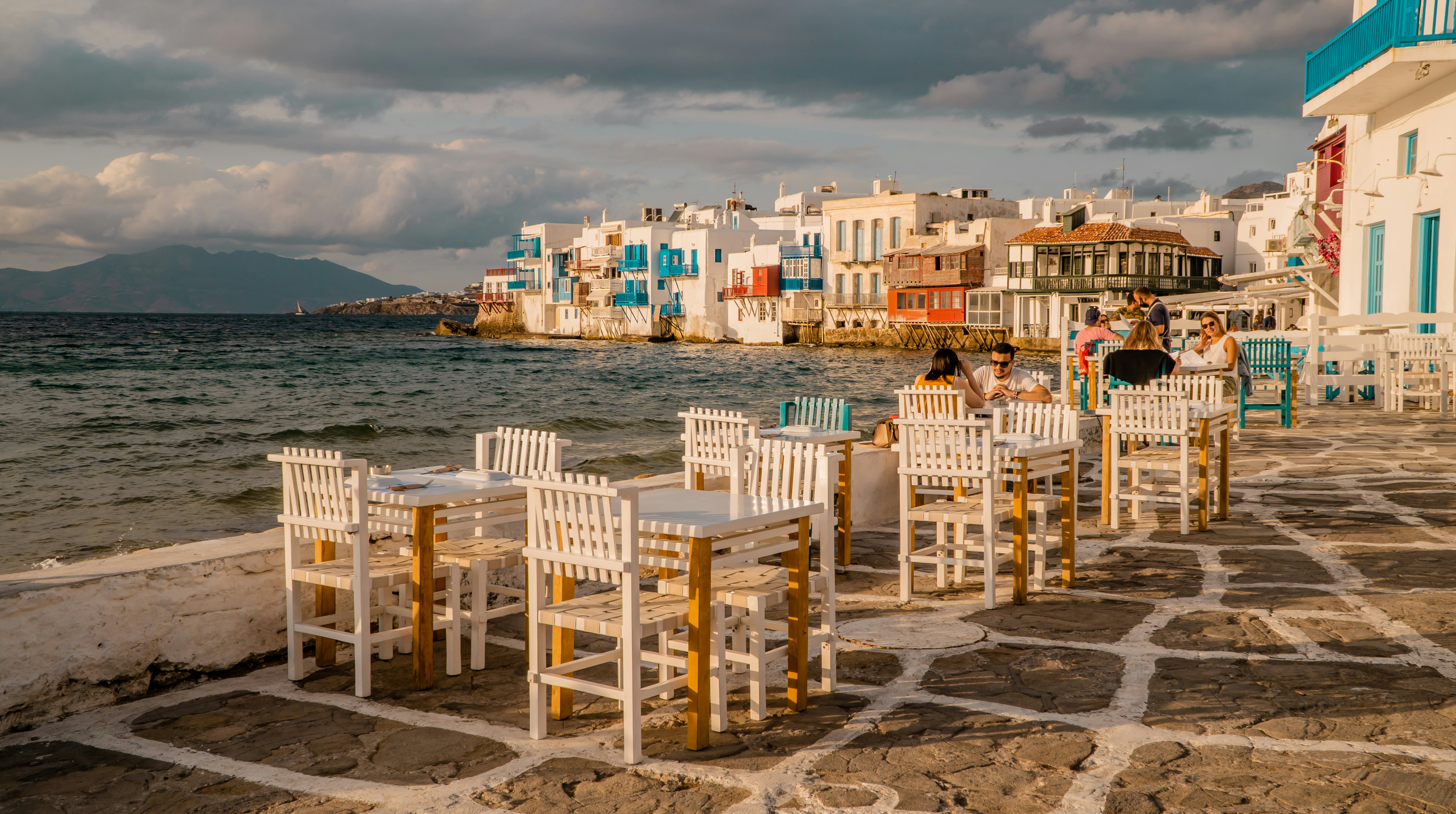 People sit on white chairs at white tables set on a walkway by the sea in Greece at sunset.
