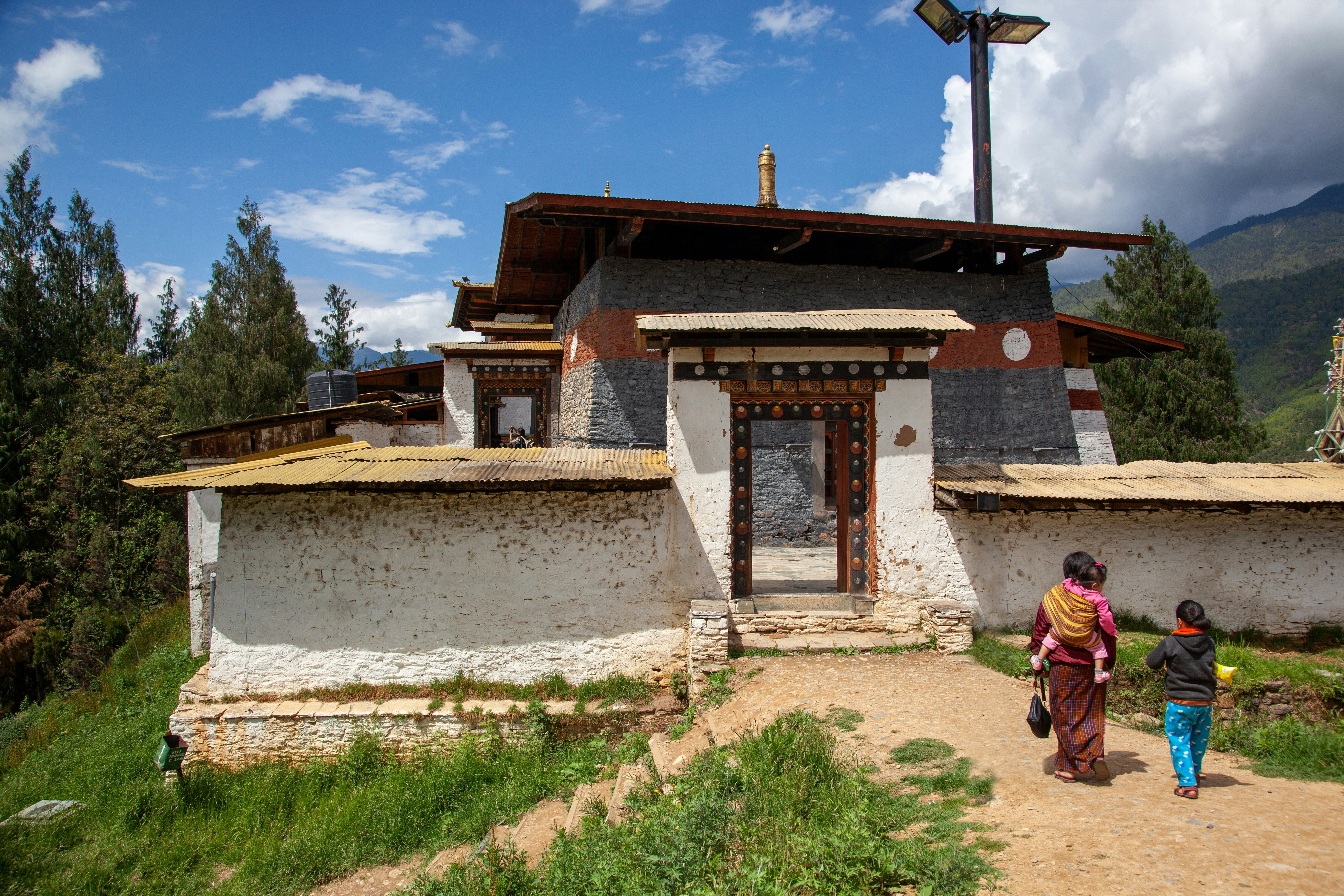 A woman with a child on her back another child beside her walks toward a low-profile building, angular, beige building with a flat roof.