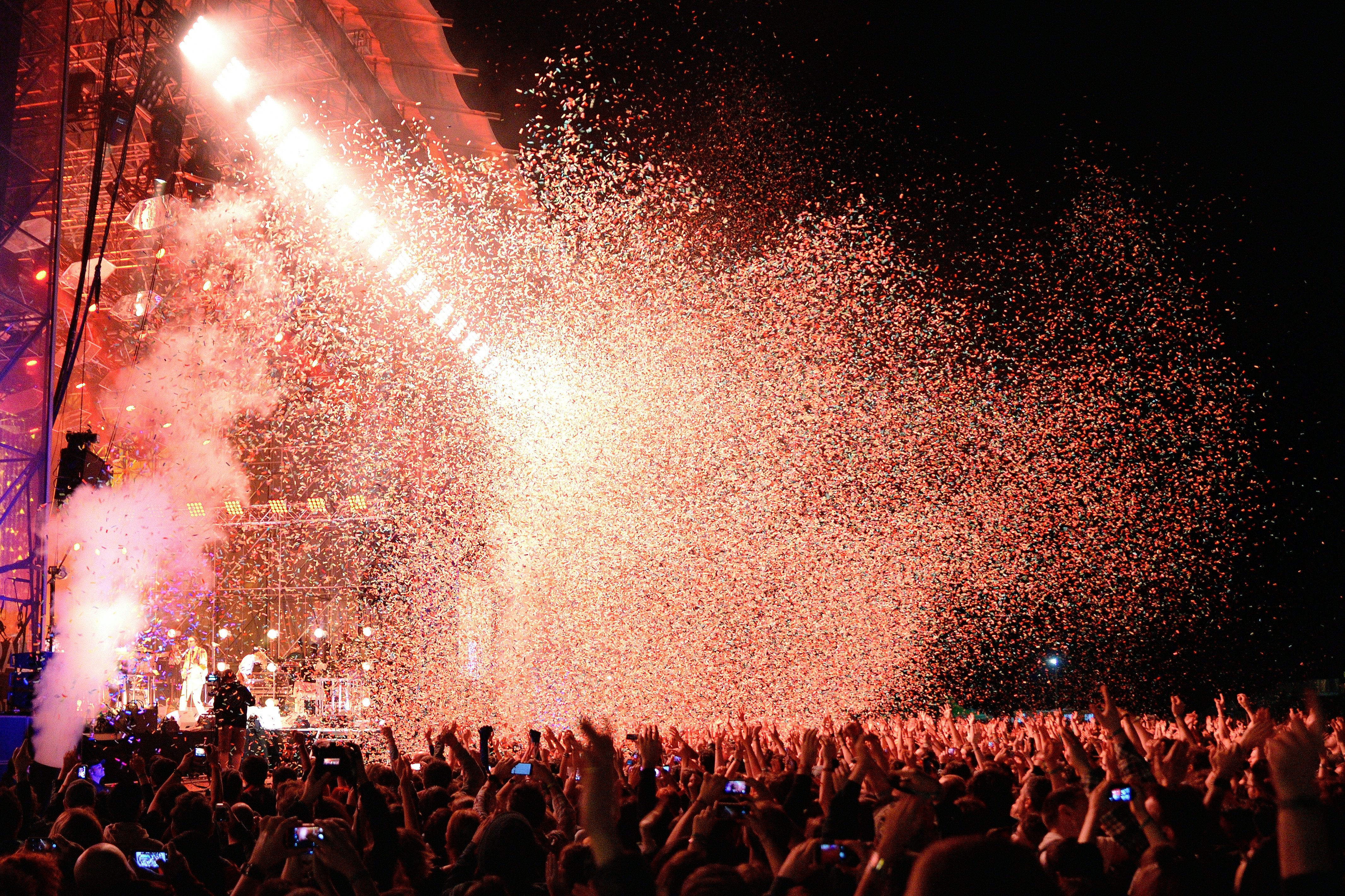 Confetti showers over a crowd of people with their hands raised in front of a lighted stage at an evening outdoor concert