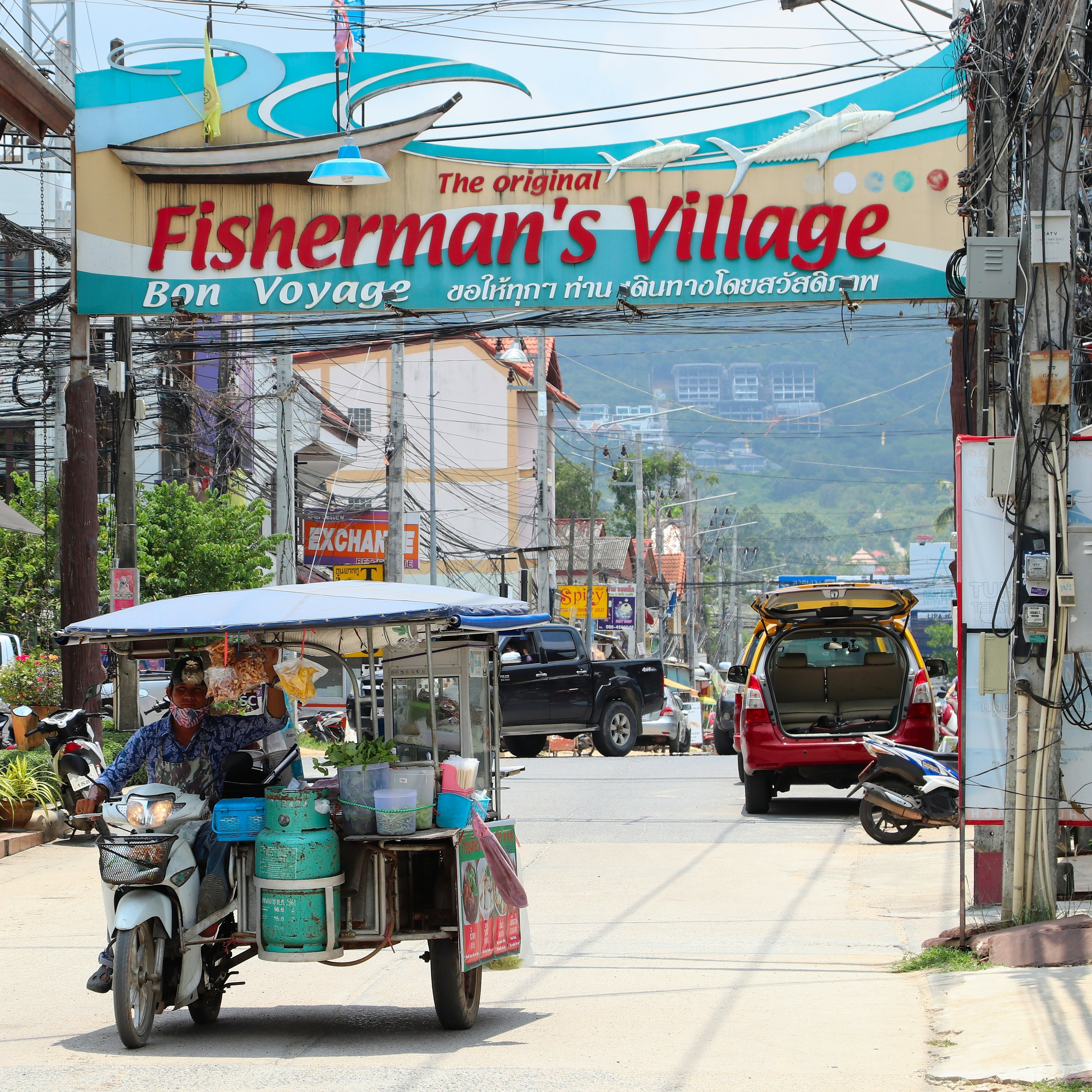 A food vendor with a cart attached to a motorcycle drives a street with a huge sign above it that says "The original Fisherman's Village, bon voyage"
