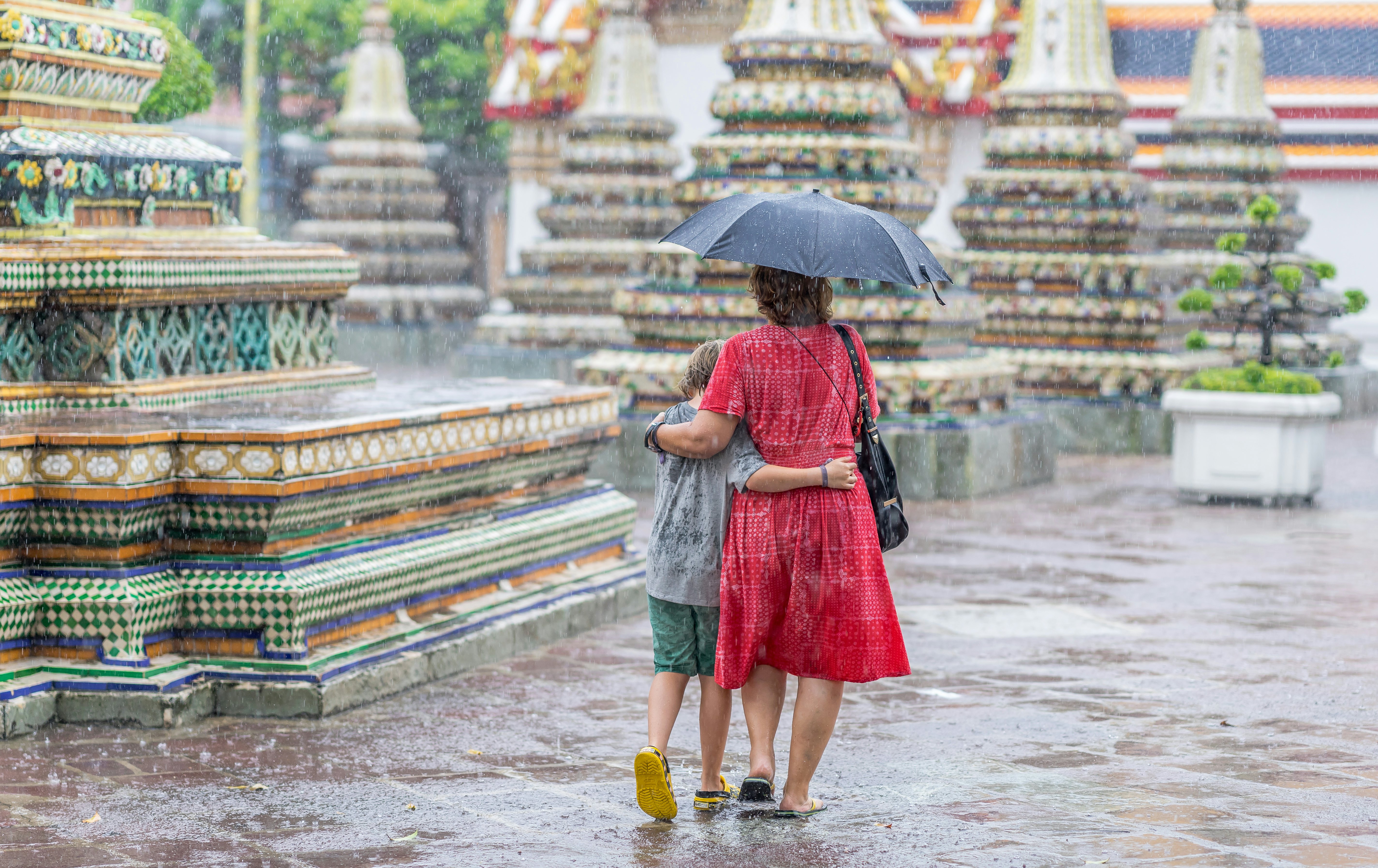 A mother and son, walking in the rain, under an umbrella at a Buddhist monastery in Bangkok, Thailand.
