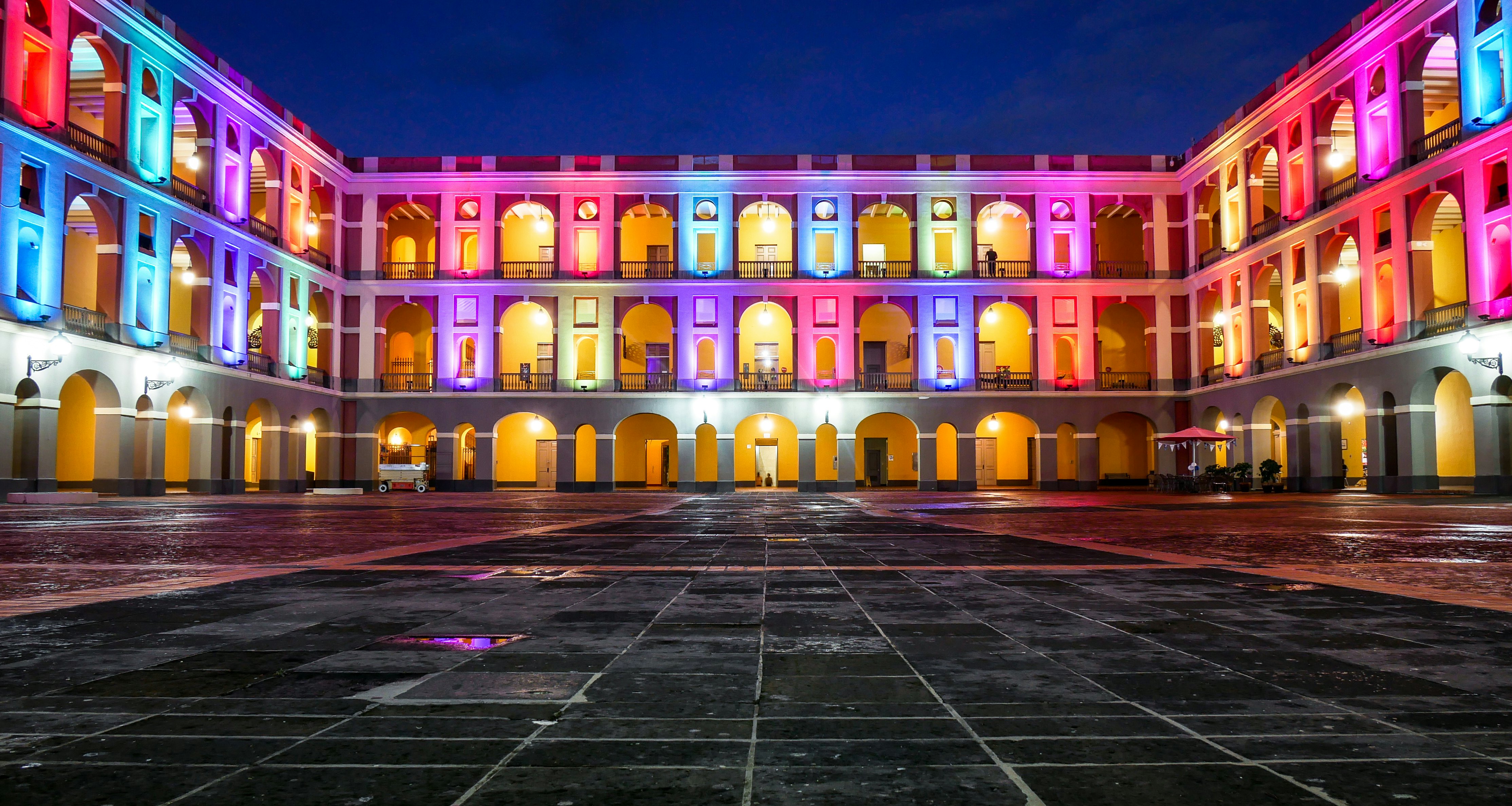 An empty courtyard between three wings of a three-story building with archways on each level. The building is lit by colorful lights at night.