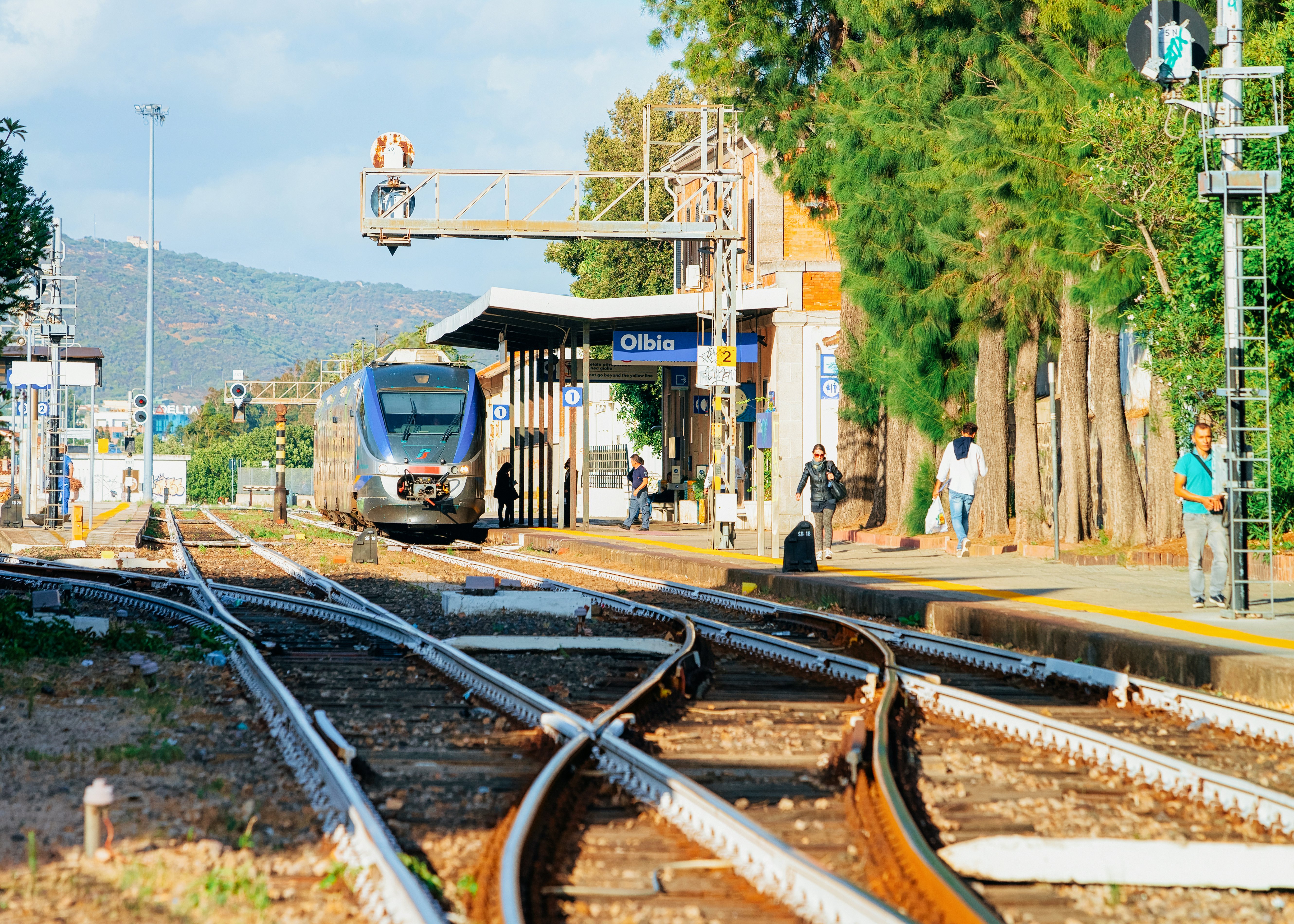 A view of tracks at a train station. People stand on a platform next to the tracks, and a stopped train is visible in the distance.