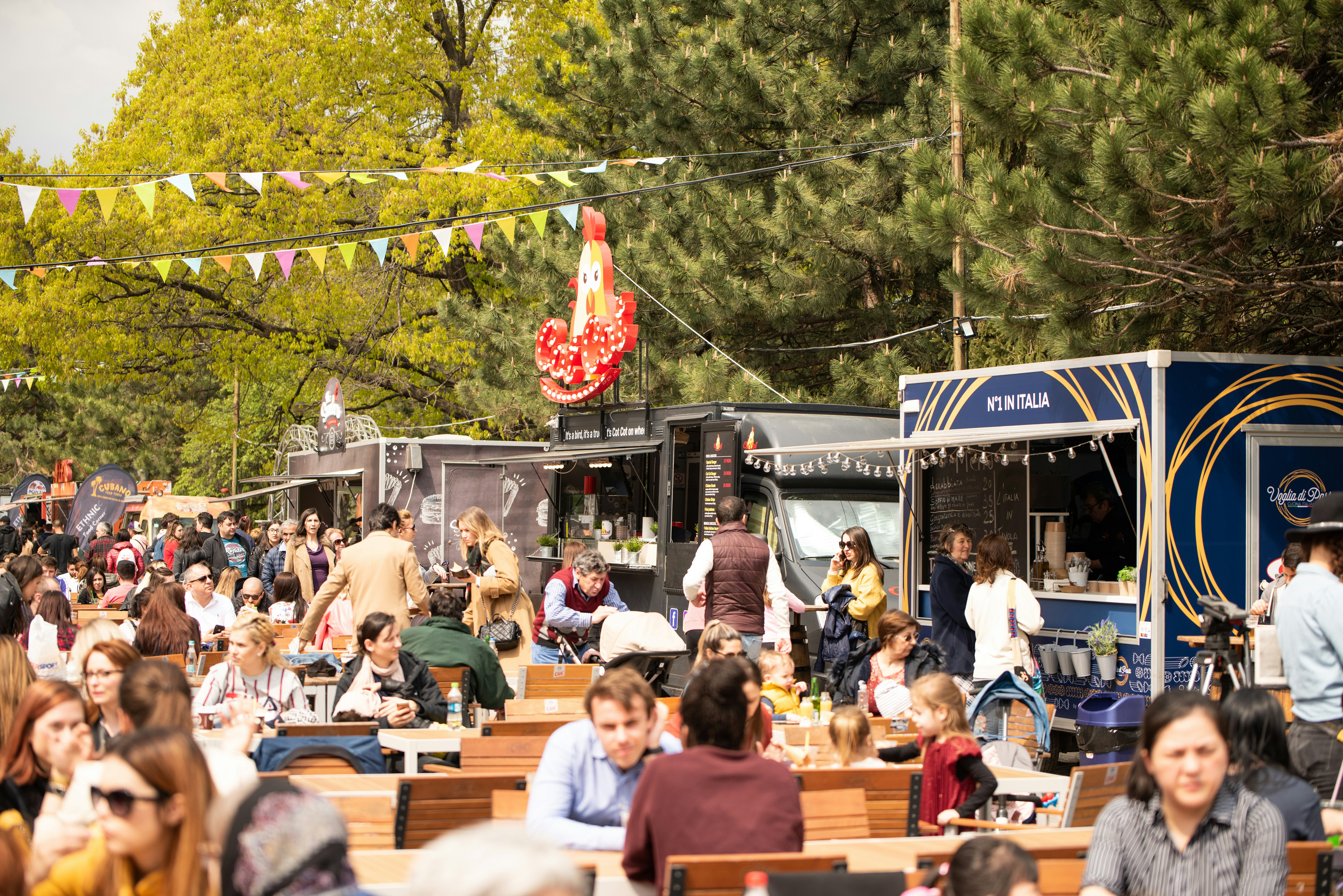 People sitting and eating at tables at a food festival in a park. Food vendors are lined up near trees in the background.