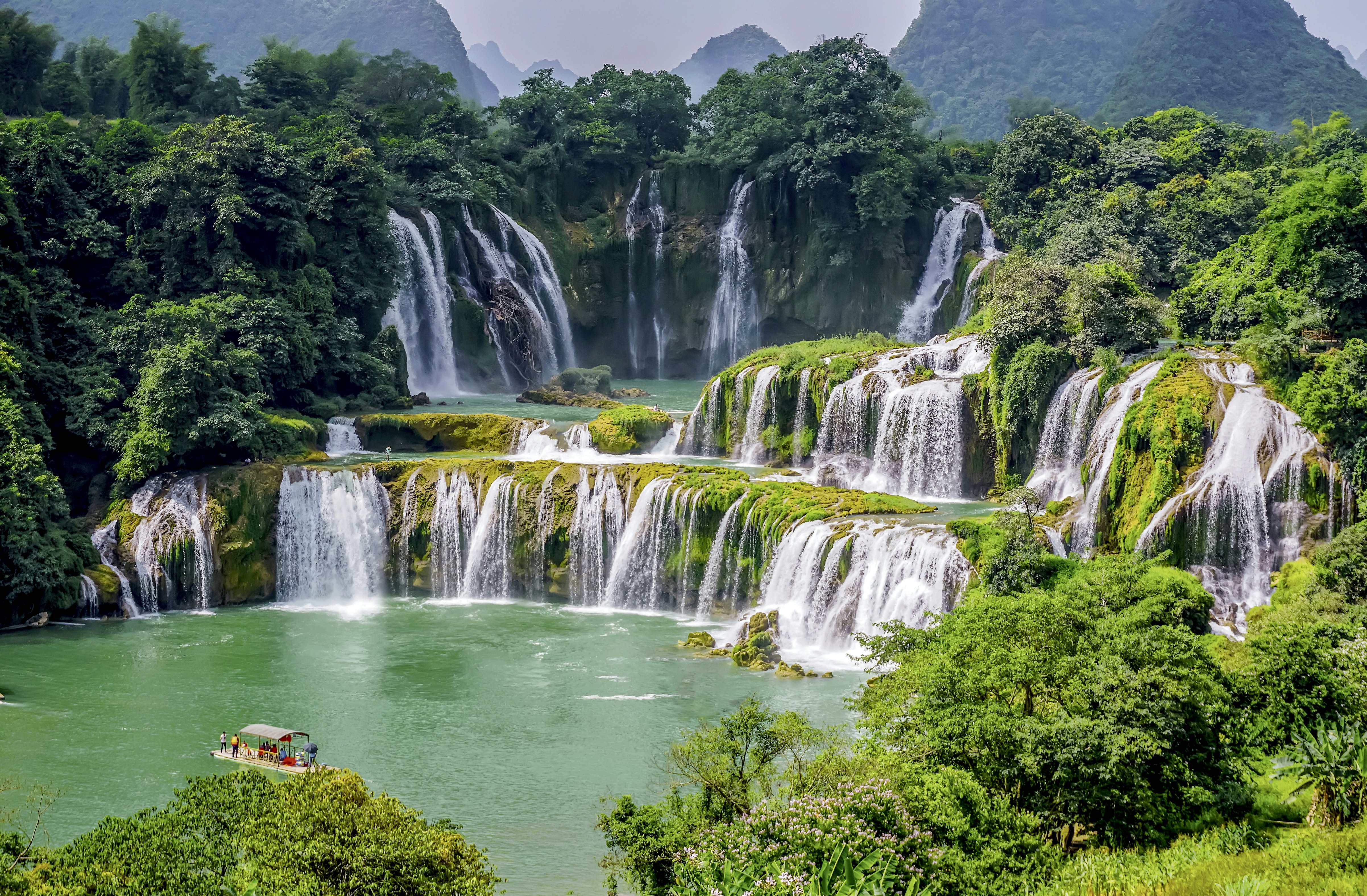 Several levels of waterfalls in Vietnam cascade out of a lush green landscape into a pool with a small viewing platform.