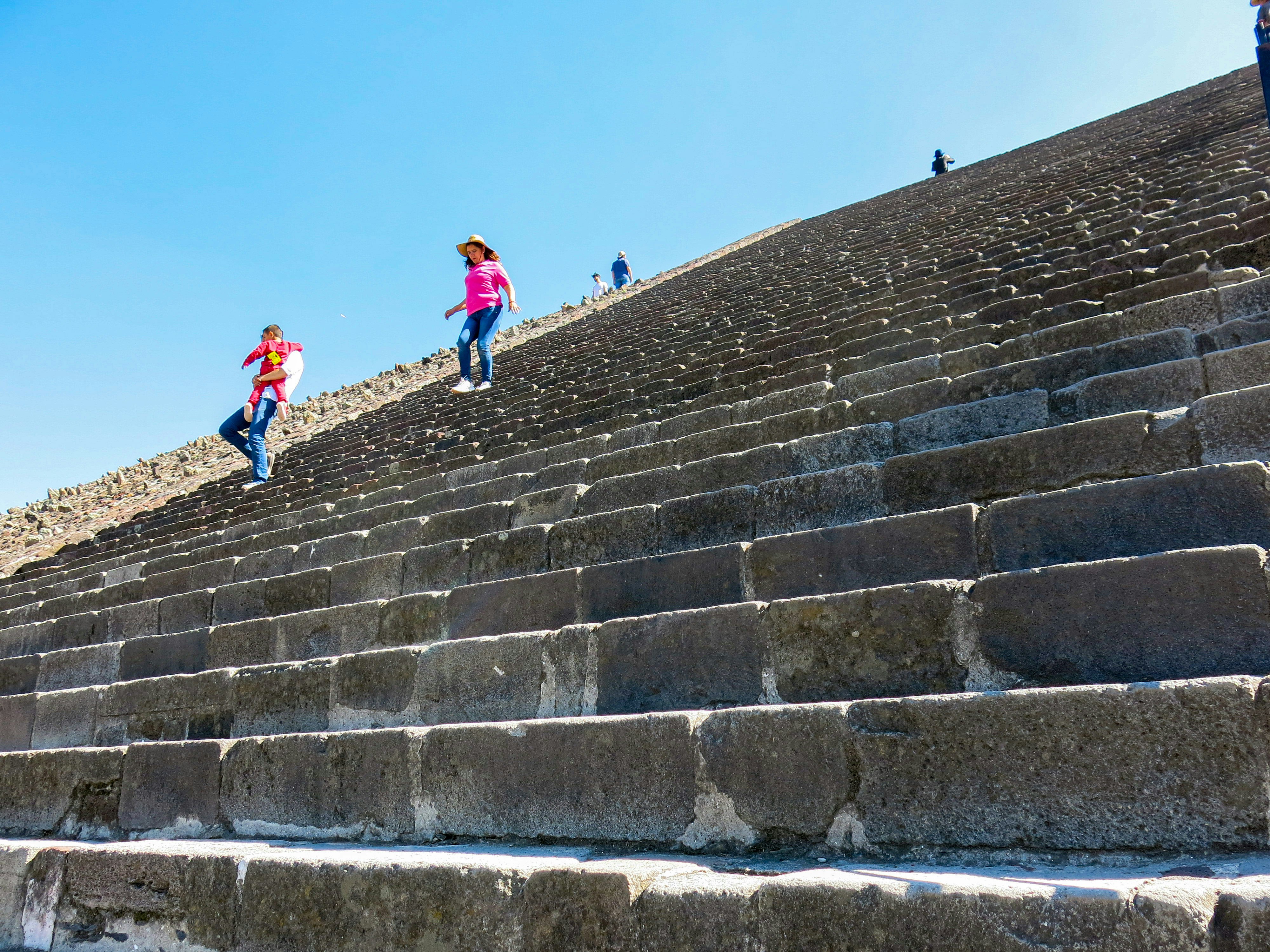A man carries a child, followed by another woman, down a set of steep steps on a pyramid.