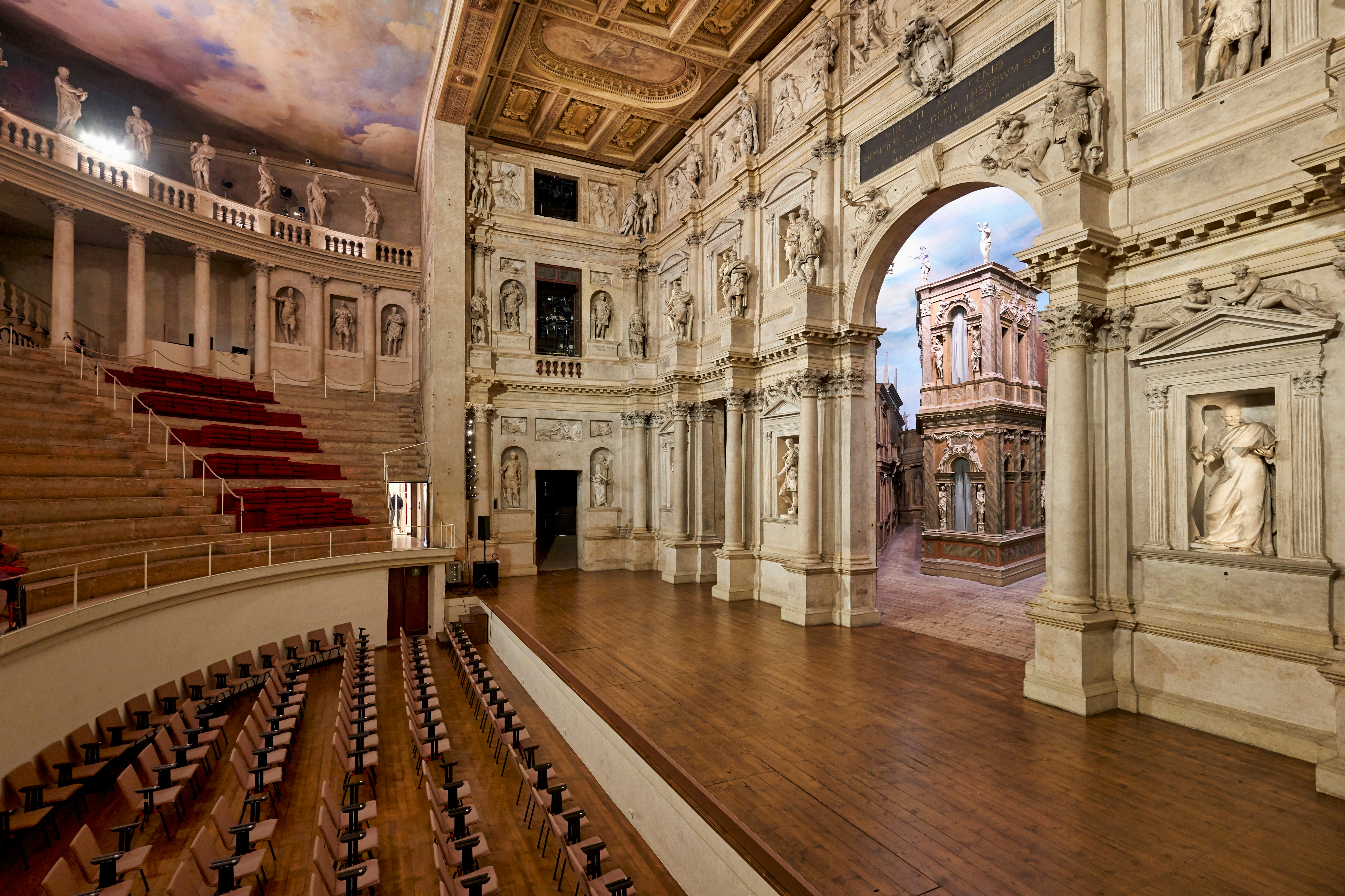 The stage of a theater in Italy has ornate carvings behind a wood stage; steeply graded benches are in the upper tier, and chairs are on the floor below the stage.