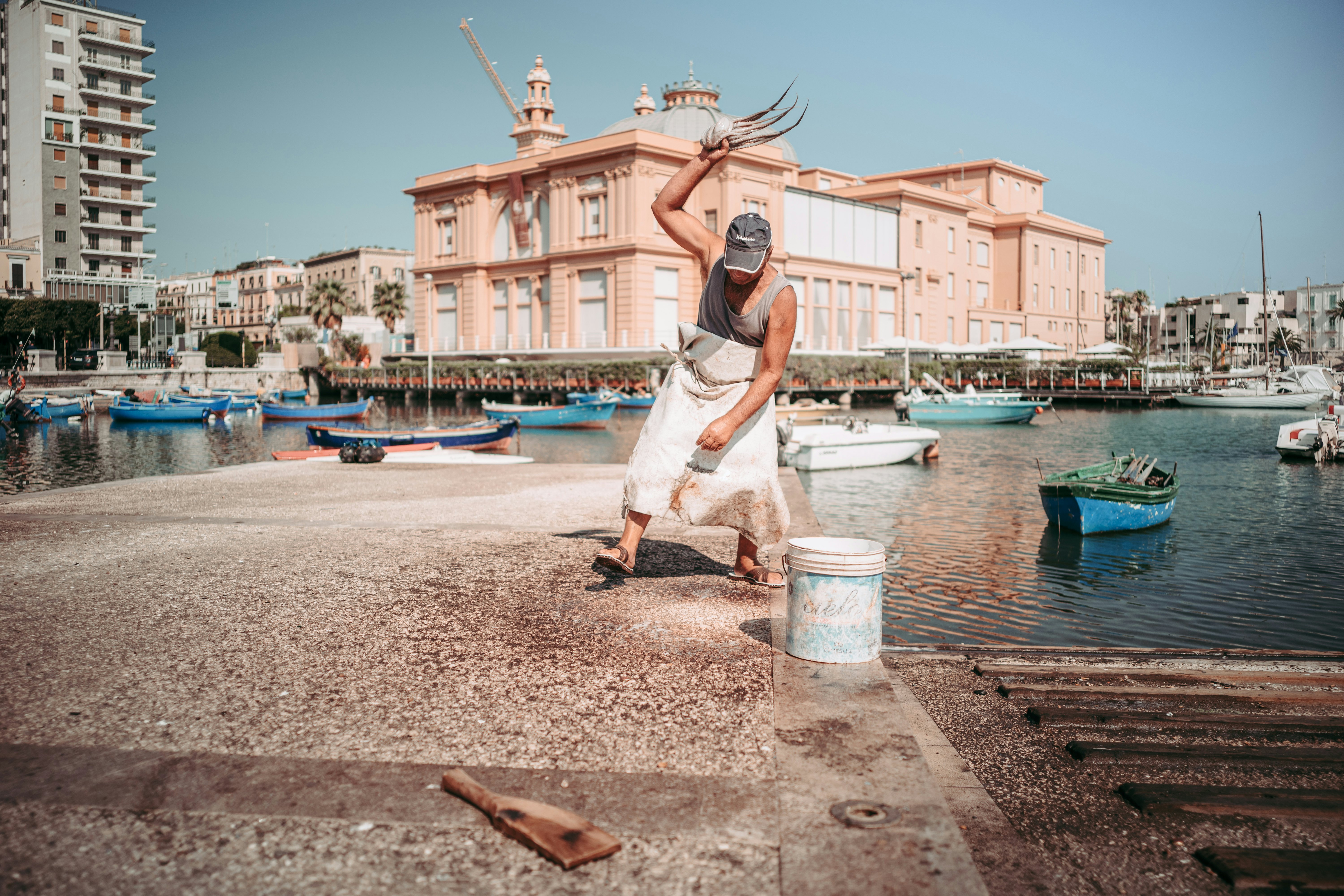 A fisherman on a pier in a city marina whips freshly caught octopus, preparing it for sale. An ornate building is seen behind him on the waterfront.