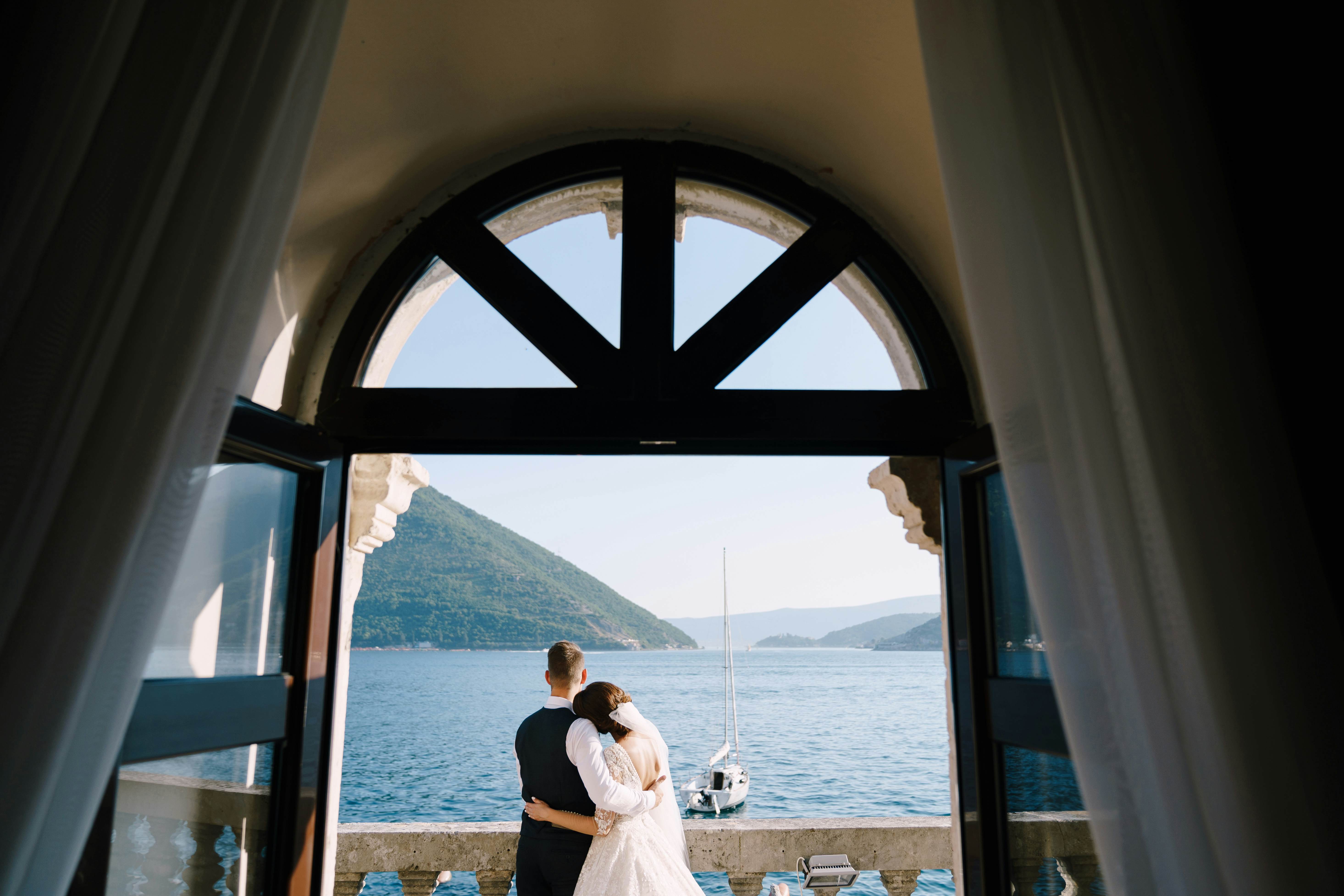 Wedding couple stand on a hotel balcony with sea view, view through an open antique window. Fine-art wedding photo in Montenegro, Perast. , License Type: media, Download Time: 2026-01-09T18:23:17.000Z, User: adouglaslott59, Editorial: false, purchase_order: 65050 - Digital Destinations and Articles, job: online editorial, client: Best places for a destination wedding, other: Ann Douglas Lott
