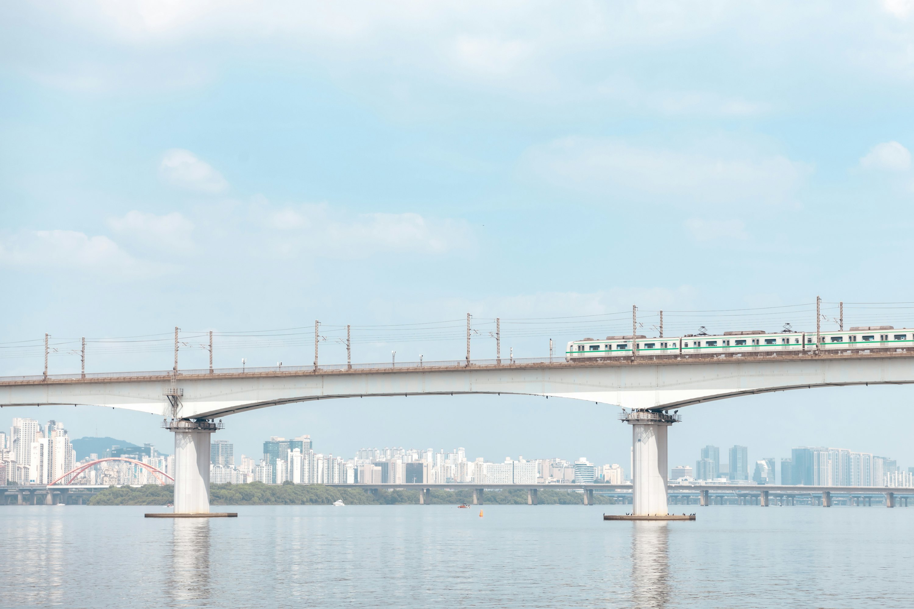 A train is seen on a bridge that crosses a wide river on a bright day. Towers of a city are seen on the shore in the distance.