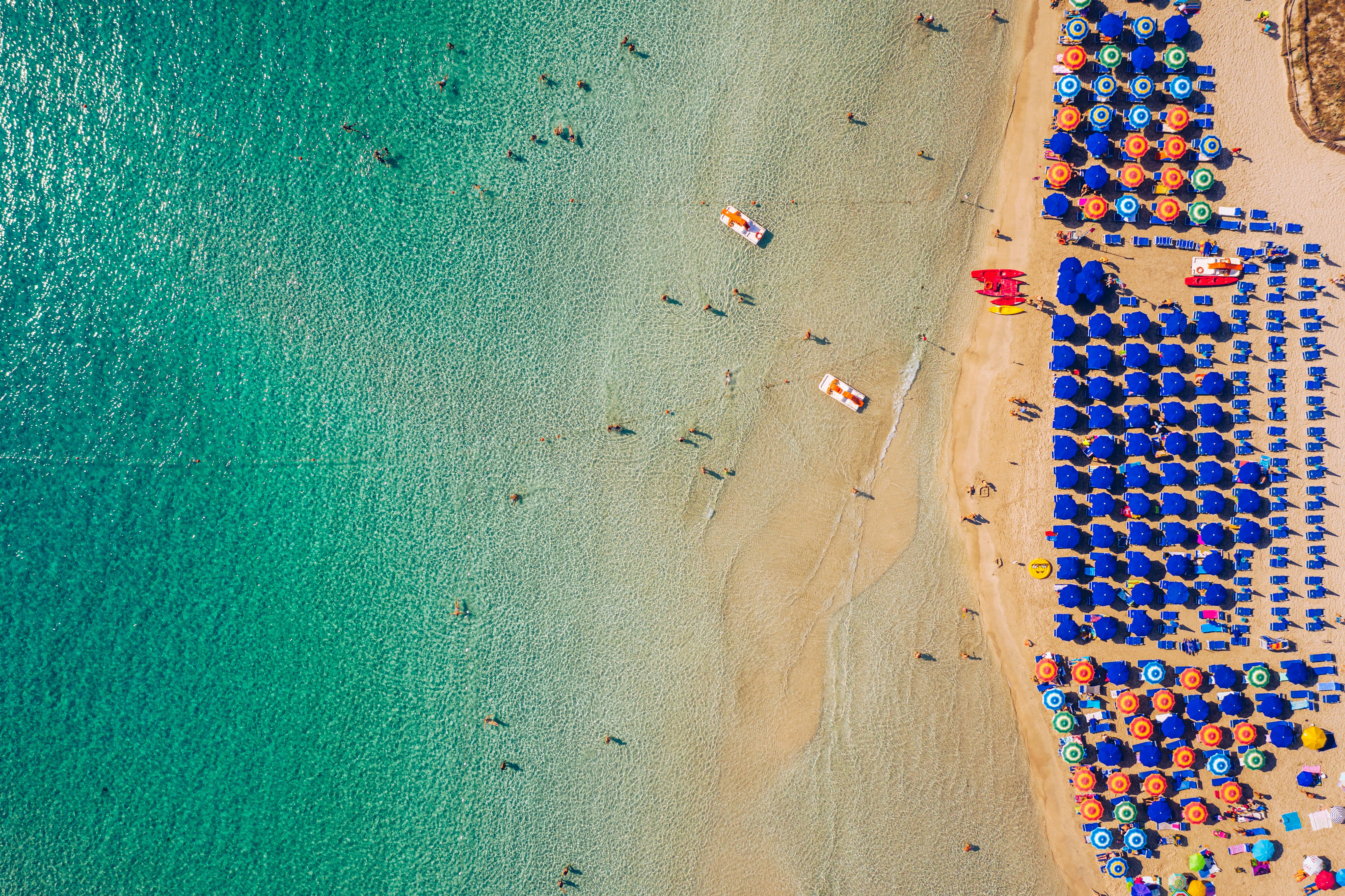 Beachgoers splashing in the shallow ocean or relaxing under sunshades on a busy beach lined with bright blue umbrellas.