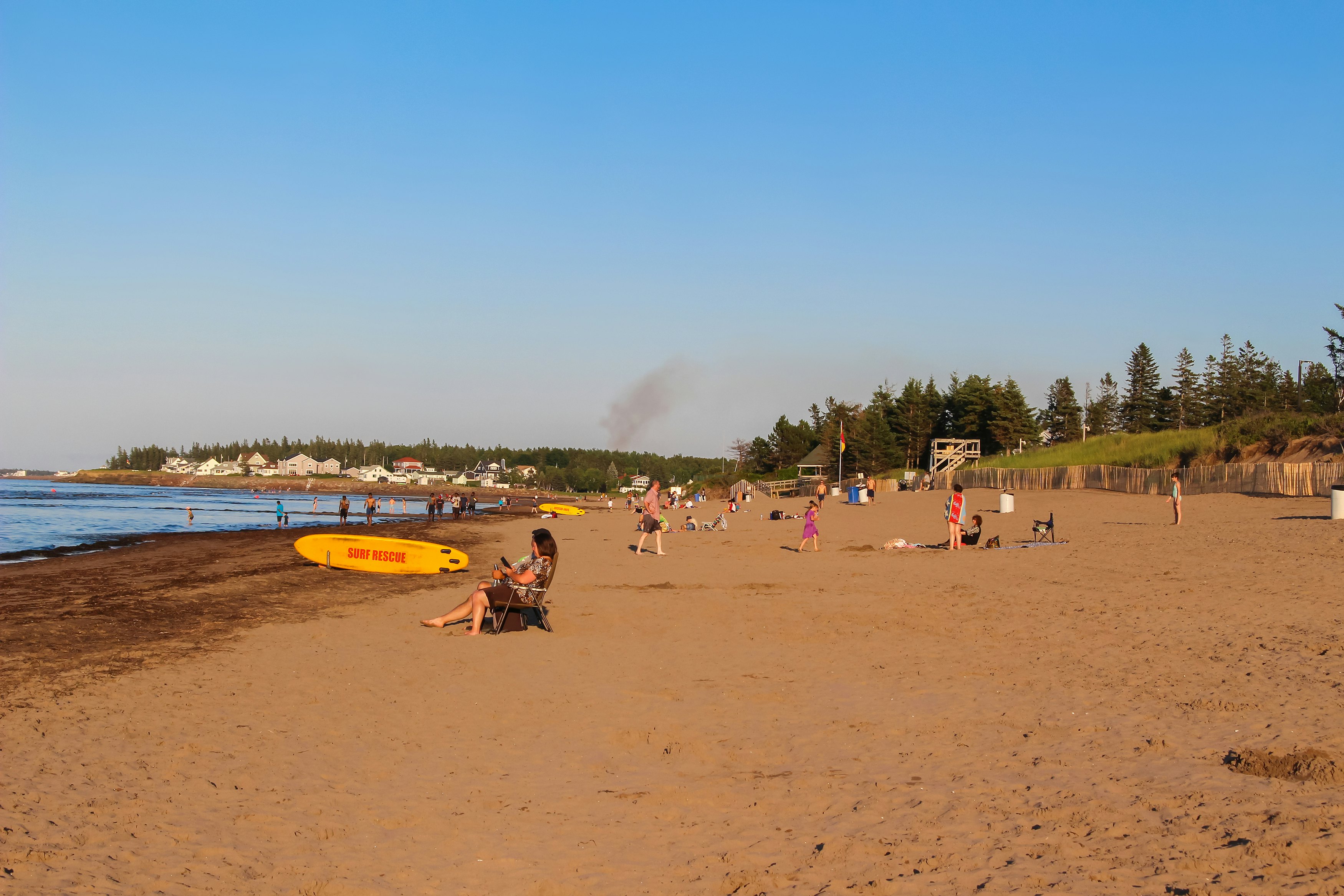 People sit on a wide beach with golden sand, with houses and businesses seen in the distance.