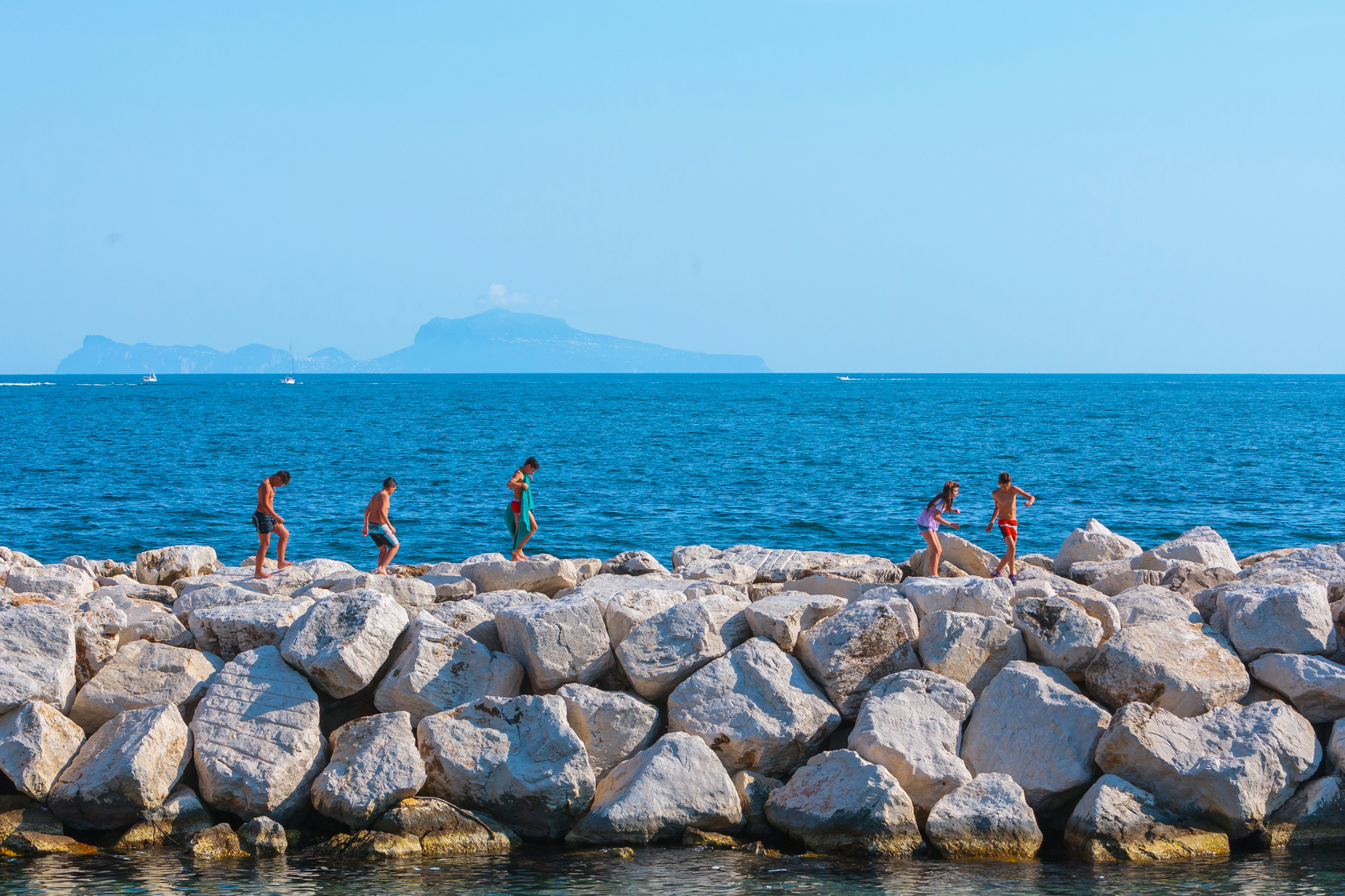 People walk along the large rocks of a breakwater next to the blue sea.