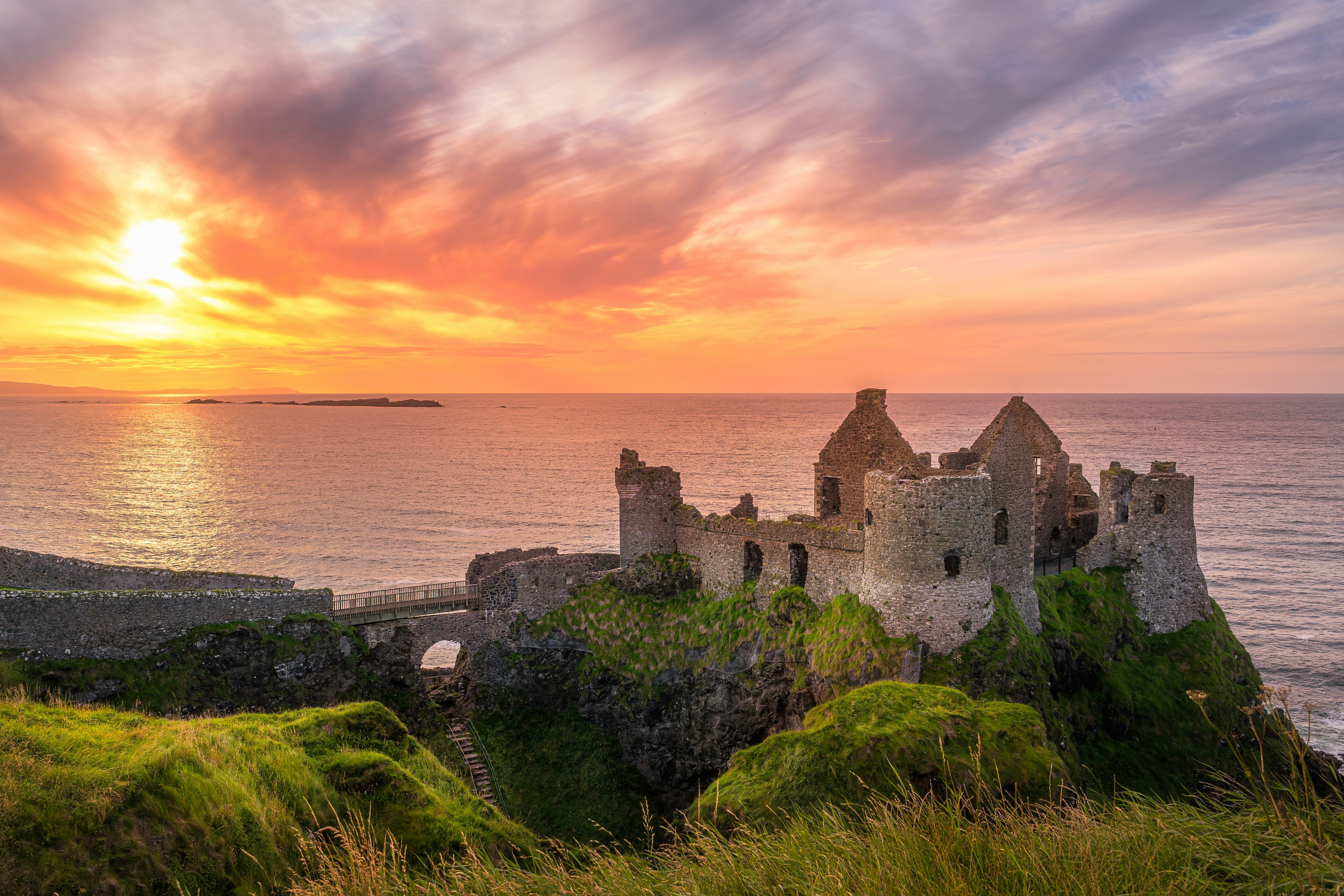 Ruins of a castle at the edge of a cliff. The sun sets in a colorful sky above it.